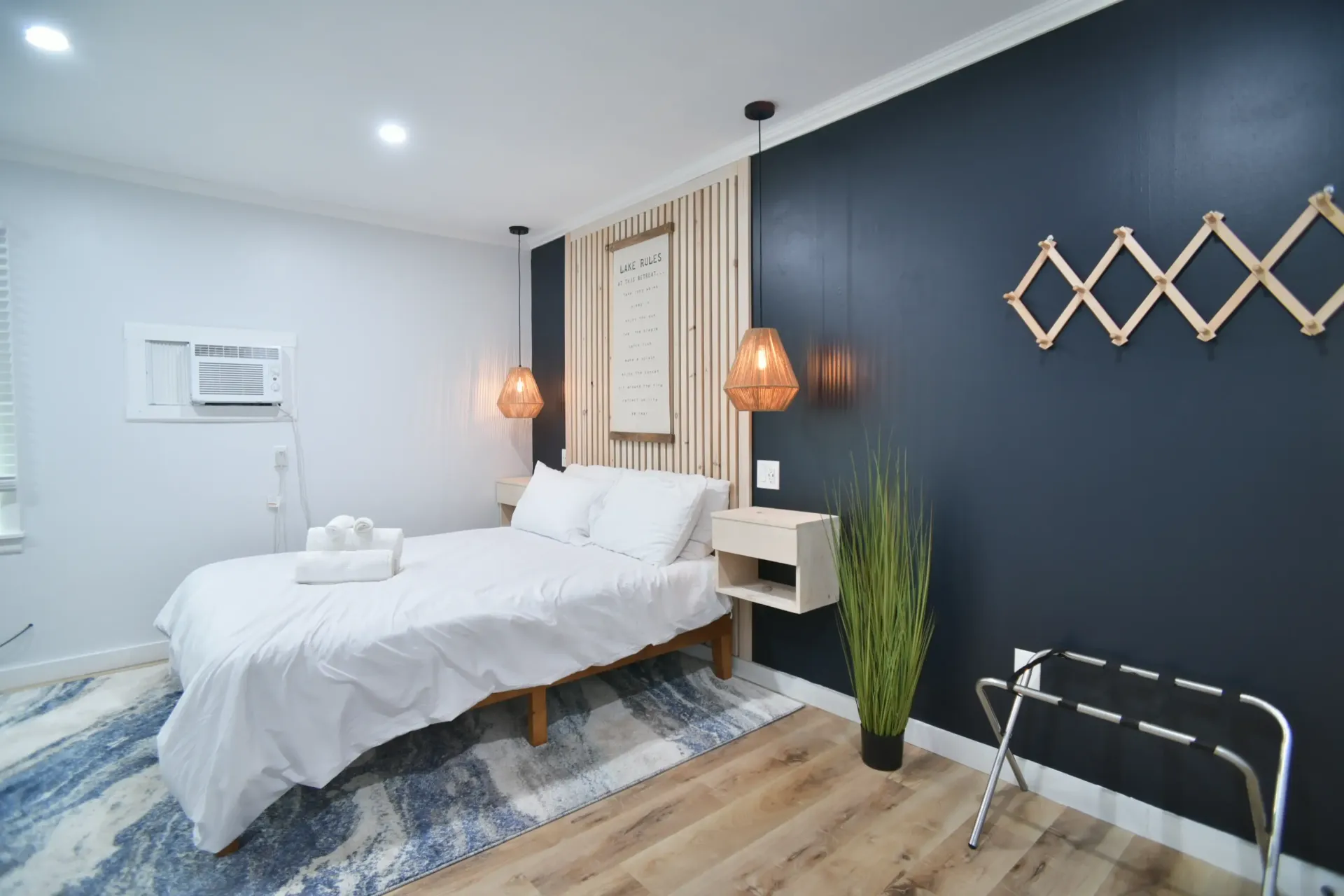 A modern bedroom with a white bed, patterned blue rug, wood paneled accent wall, dark navy wall, and hanging lights.