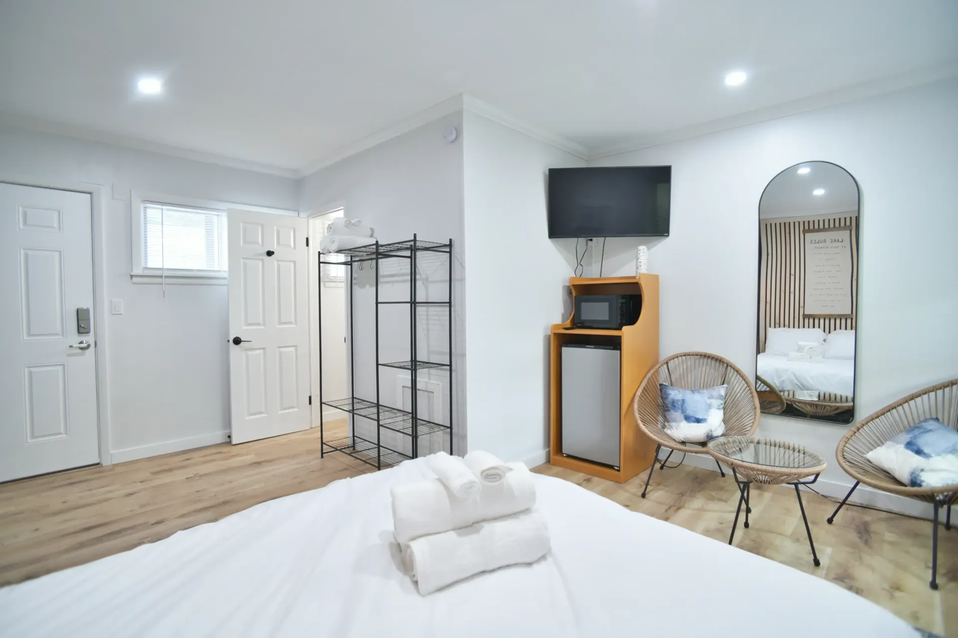 A light-toned hotel room featuring a bed with folded white towels, a metal clothing rack, a TV, and two wicker chairs.