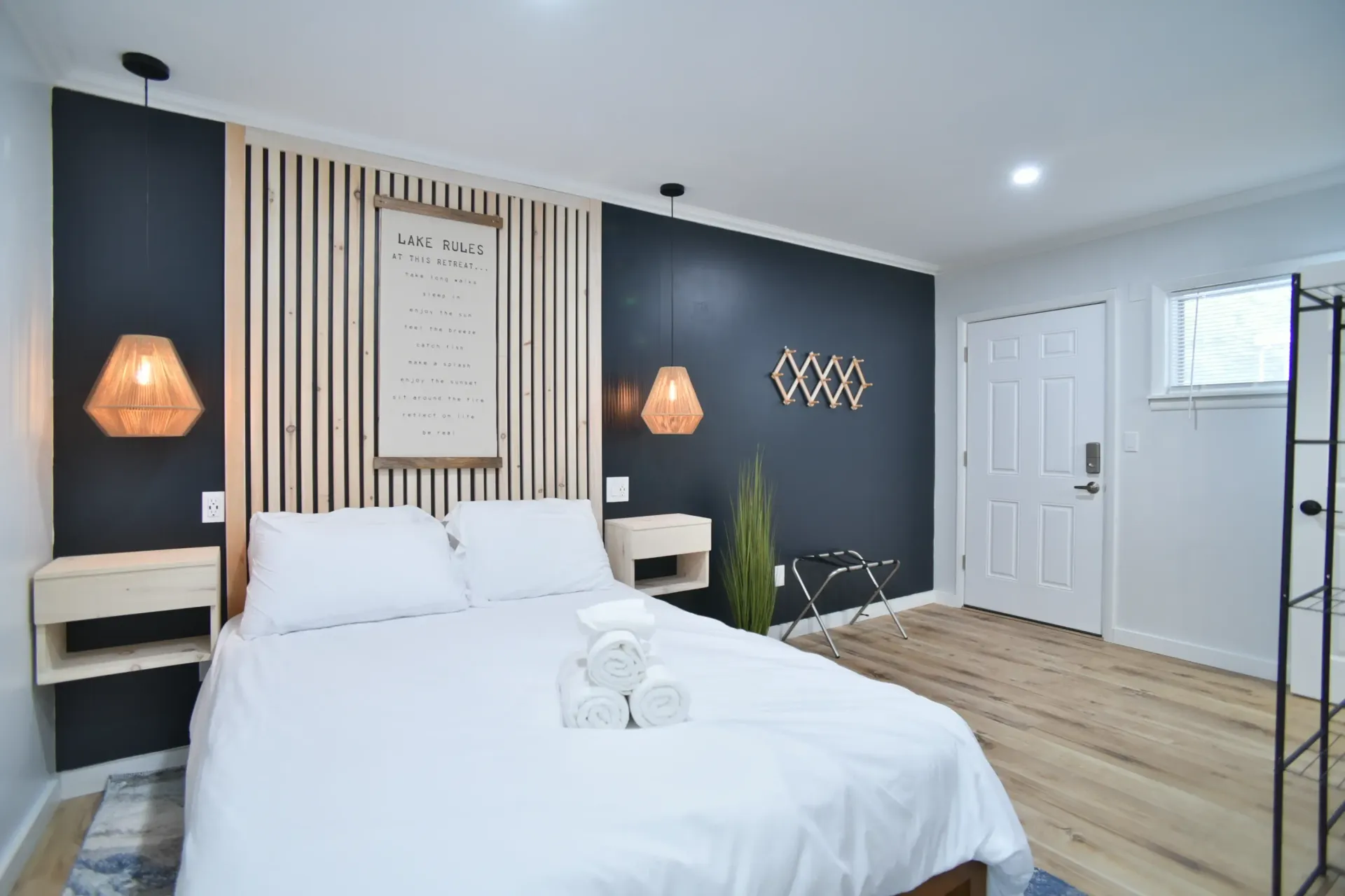 A hotel room with a queen bed, wooden slat headboard, dark accent wall, hanging pendant lights, and a white entry door.