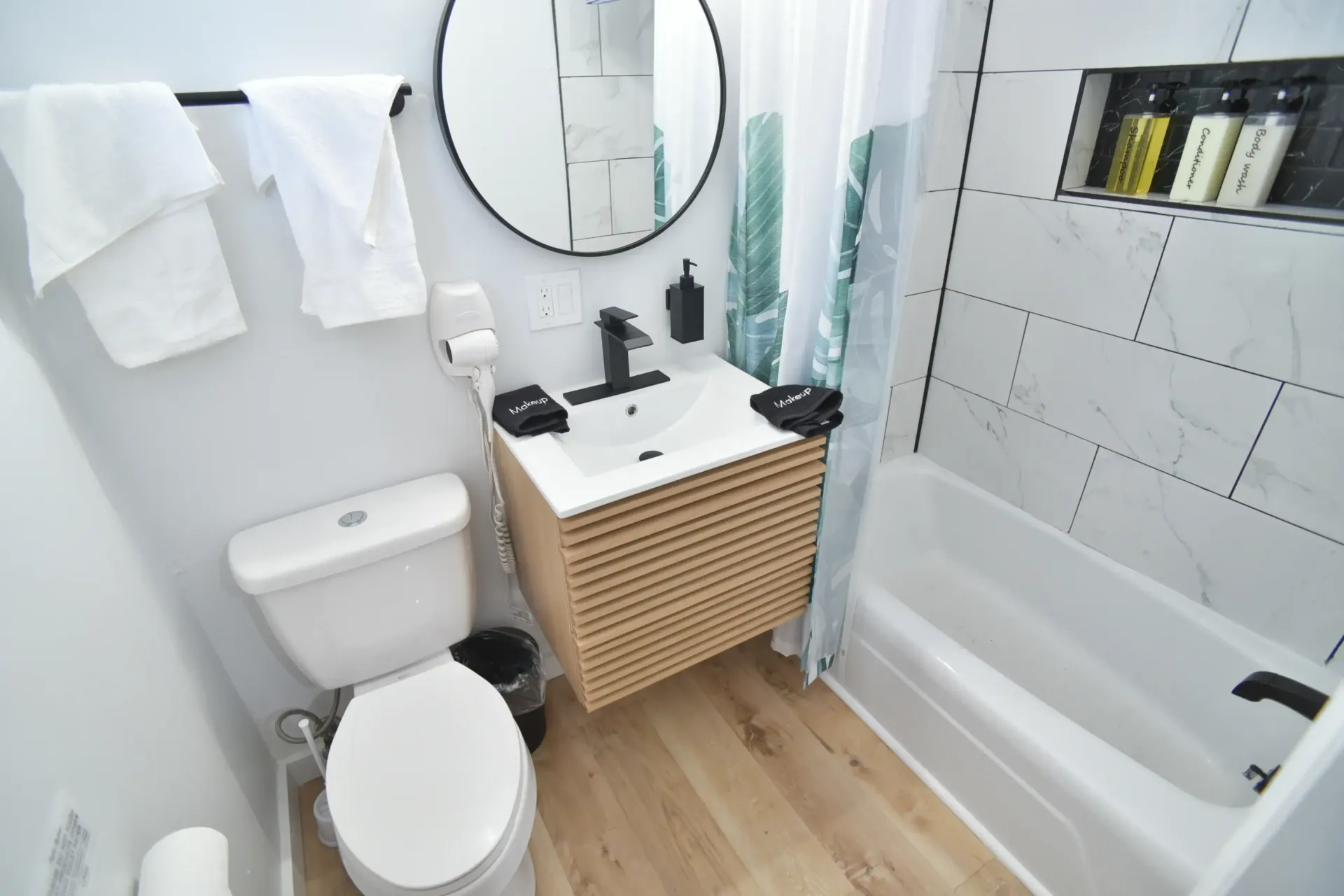 A modern, compact bathroom featuring a white toilet, a slatted wood floating vanity, a round mirror, and a tiled tub.