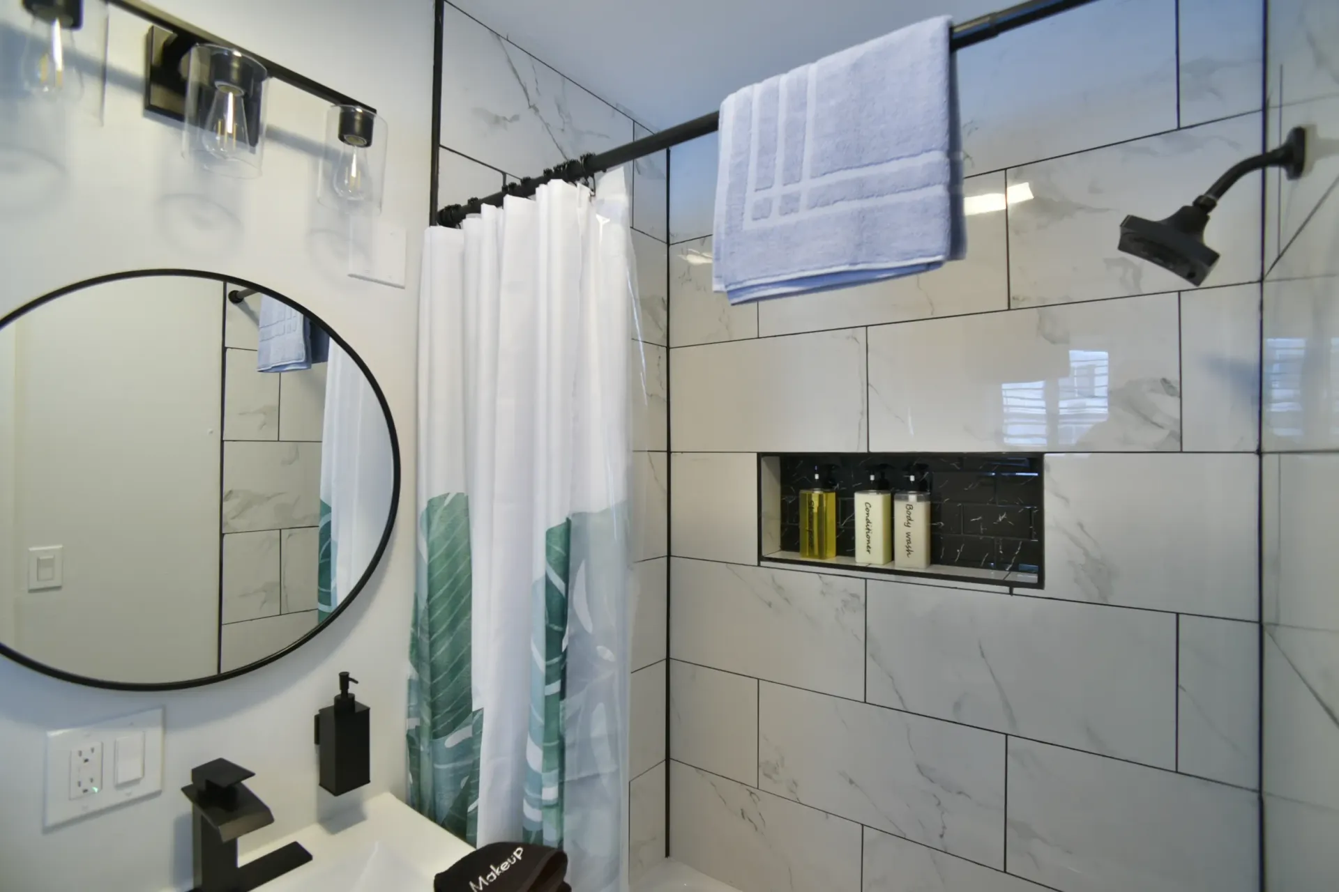 A modern bathroom featuring marble-tiled walls, a round mirror, a botanical-print shower curtain, and a black showerhead.