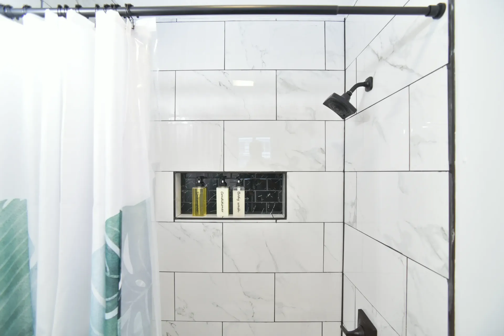 A modern shower with white marble-patterned subway tiles, a black showerhead, and a recessed niche holding three bottles.