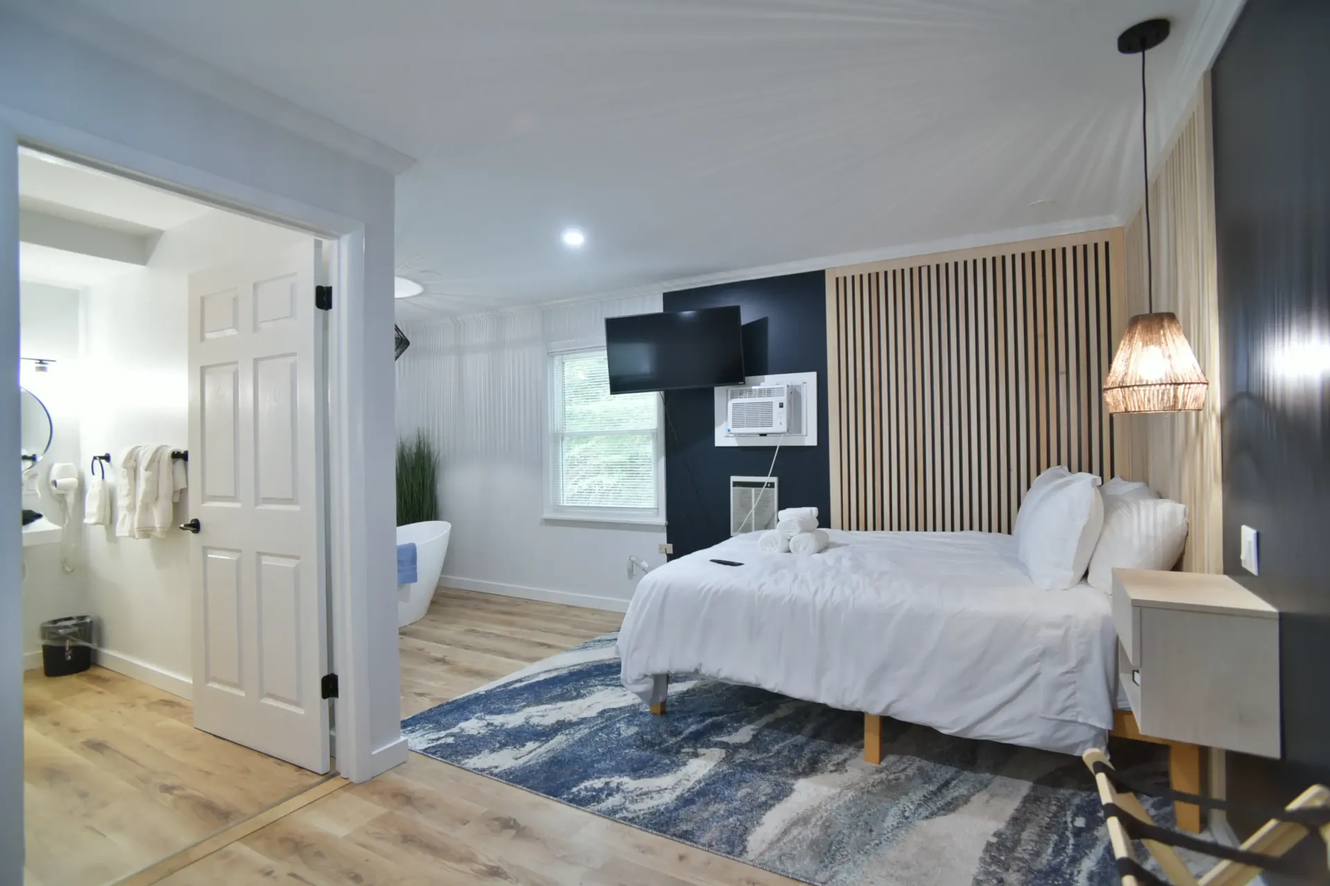A modern hotel bedroom with a queen bed, wood floors, a blue area rug, and a freestanding tub visible through a doorway.