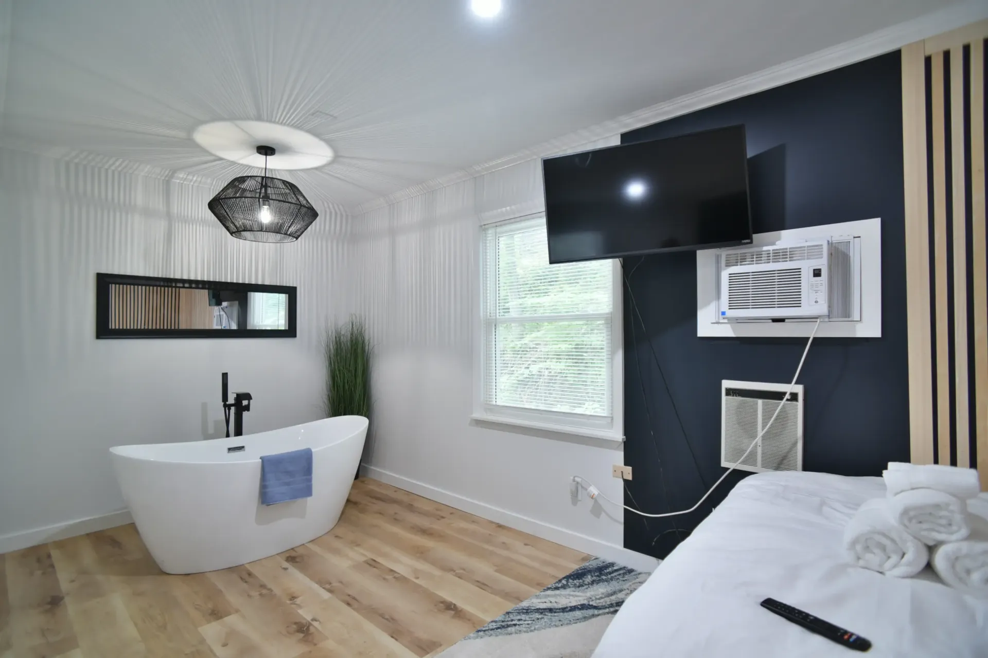 A modern bedroom featuring a freestanding white bathtub, a flat-screen TV on a dark accent wall, and a bed with white linens.
