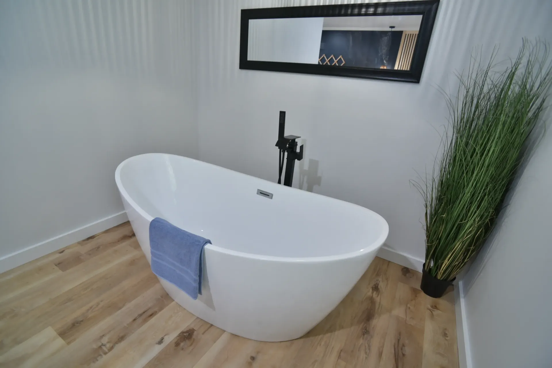 A white oval freestanding bathtub with a blue towel on its edge, next to a black floor faucet and a tall floor plant.