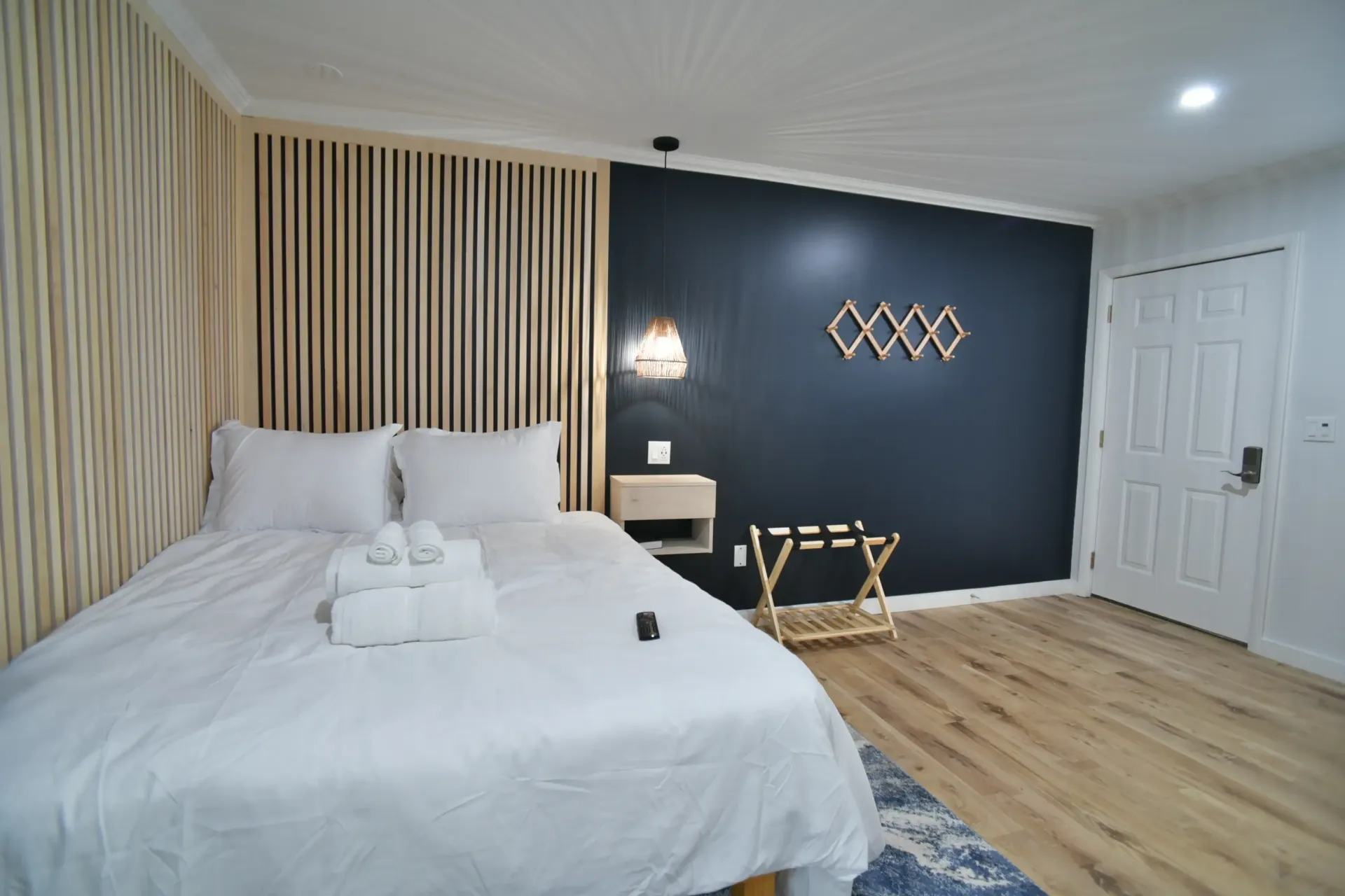 A modern hotel room with a white bed, wood-slat headboard, dark blue accent wall, wall-mounted rack, and light wood floors.