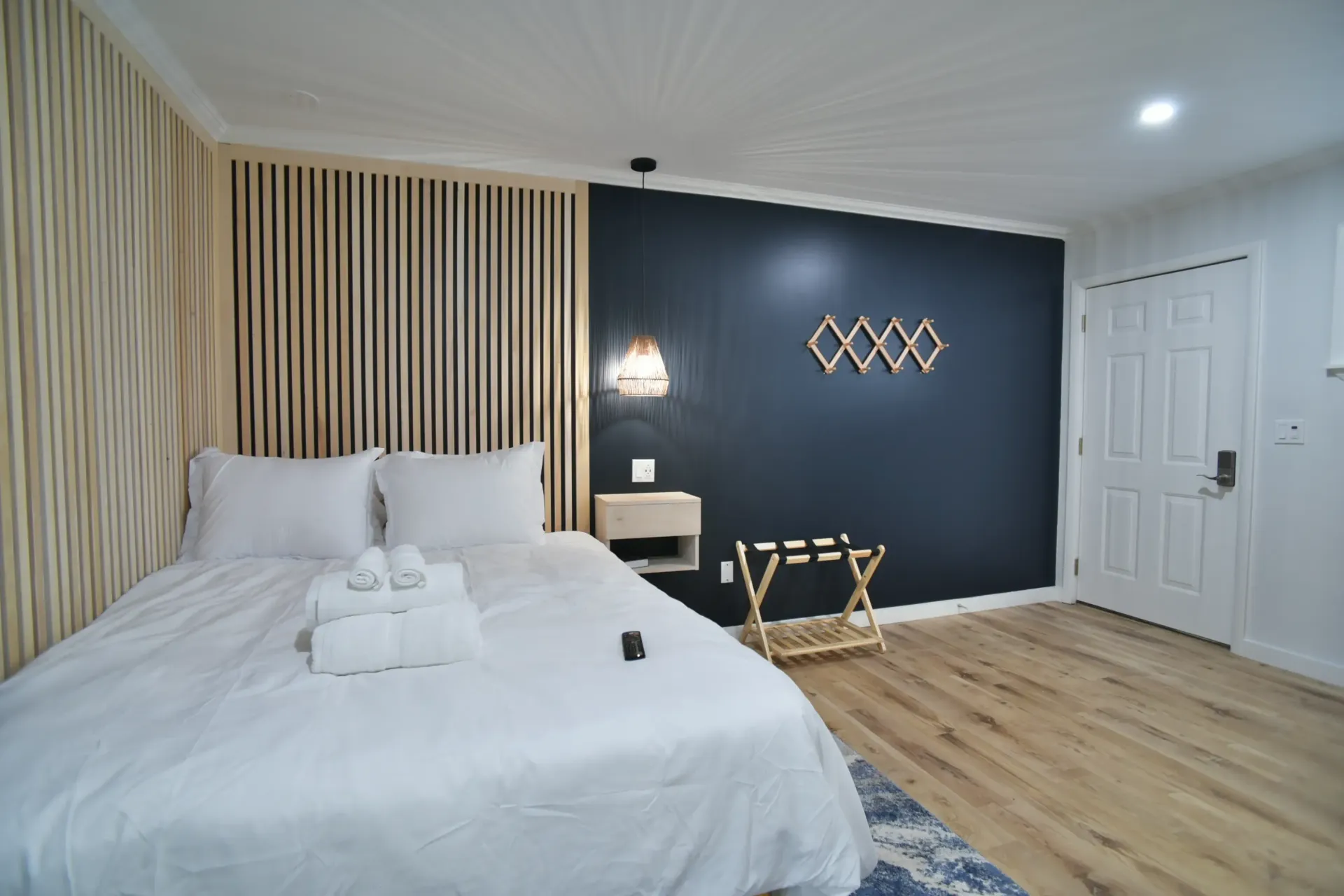 A bedroom with a queen bed, white linens, a dark blue feature wall, wood-slat headboard, and light wood flooring.