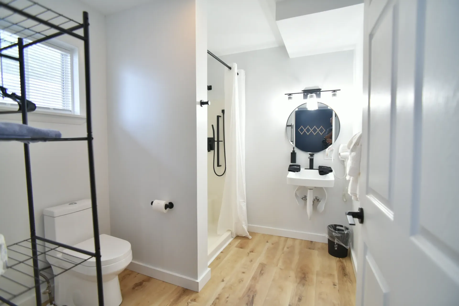 A bathroom with light-toned wood floors, white walls, a toilet, a shower with a white curtain, and a wall-mounted sink.