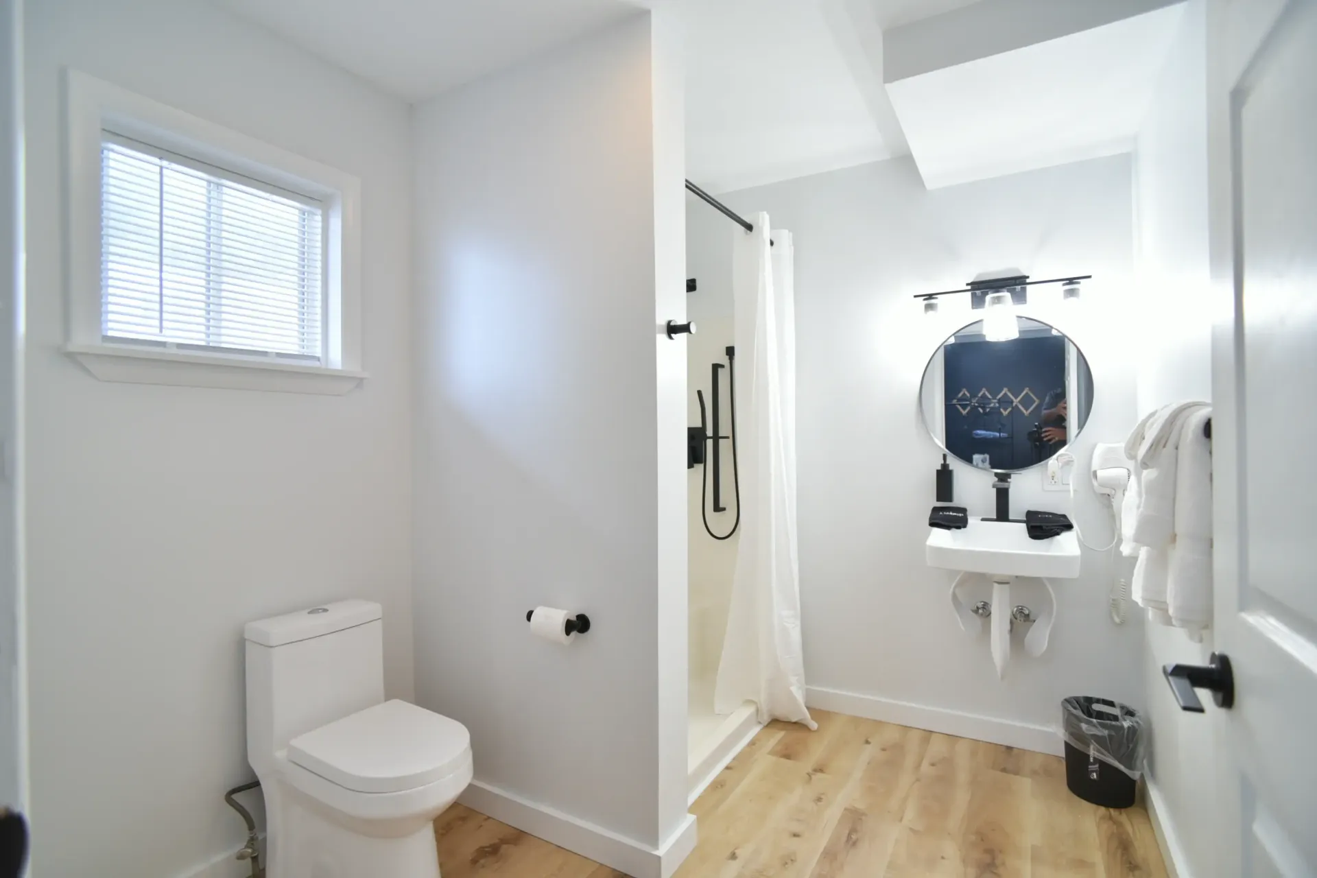 A bright, modern bathroom with a white toilet, wall-mounted sink, round mirror, and a walk-in shower with a white curtain.