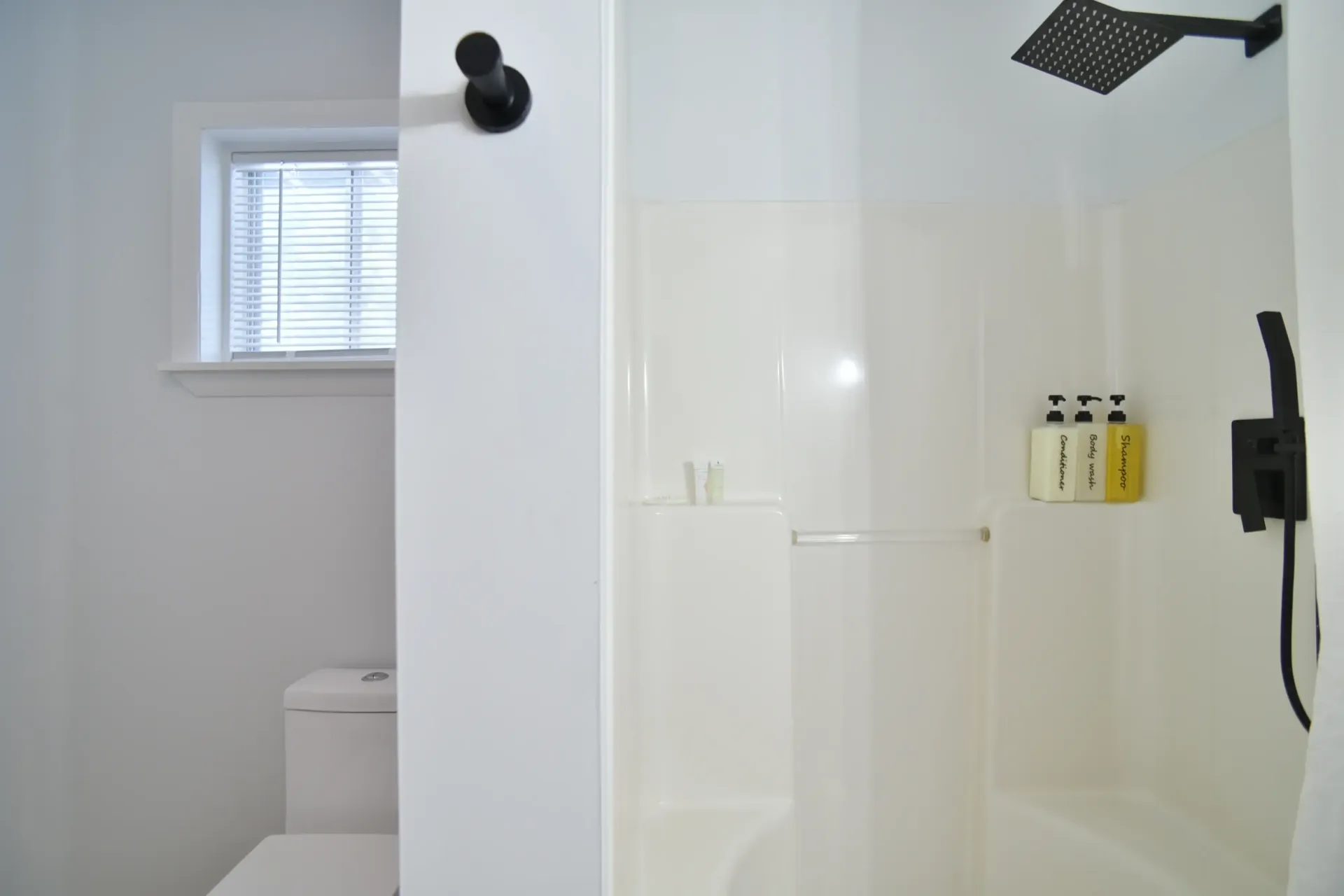 A bathroom with a white toilet near a window and a shower stall featuring a matte black rainfall head and wall dispensers.
