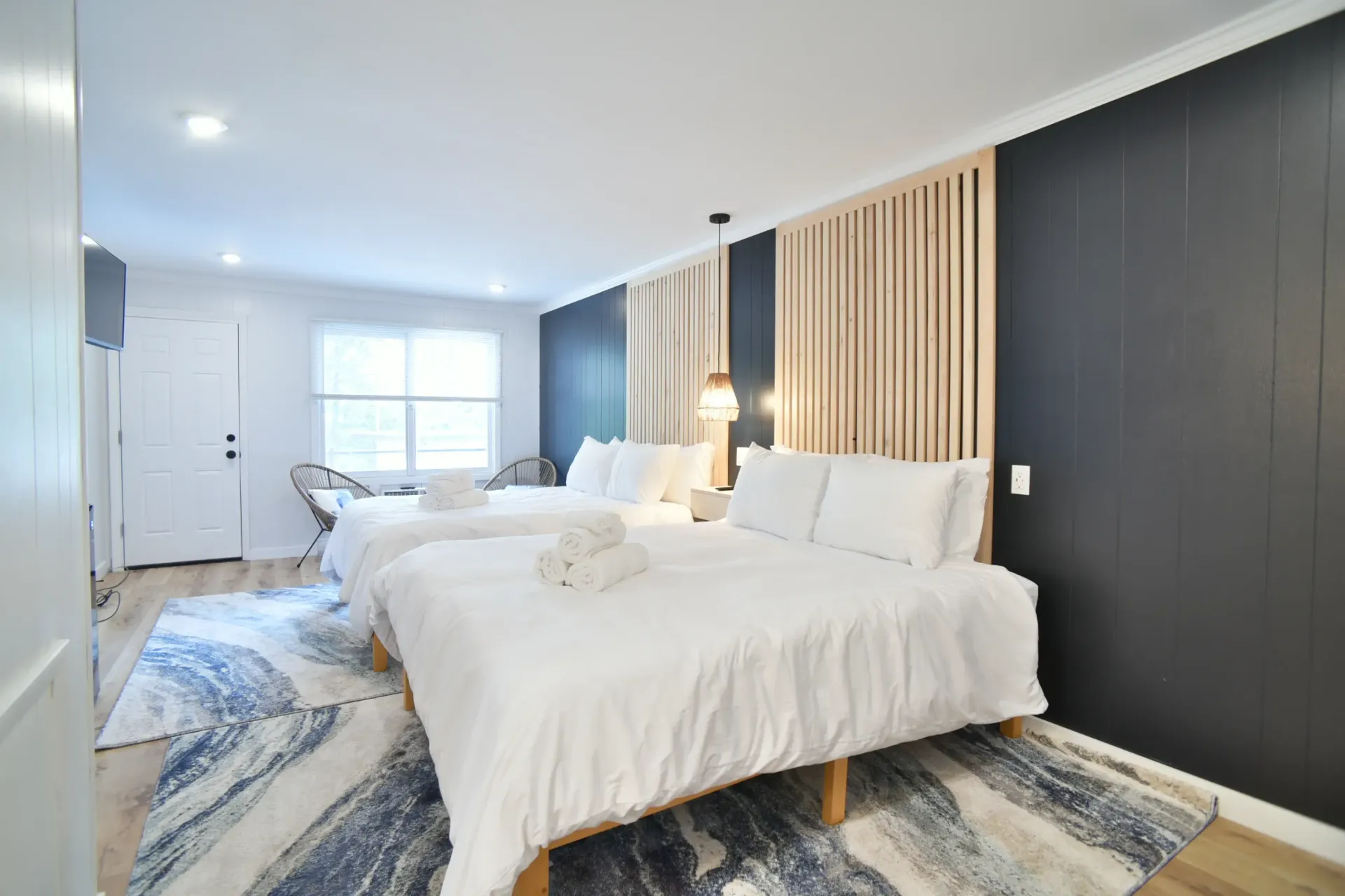 A modern hotel room with two white queen beds, a blue and white patterned rug, and a dark accent wall with wood slats.
