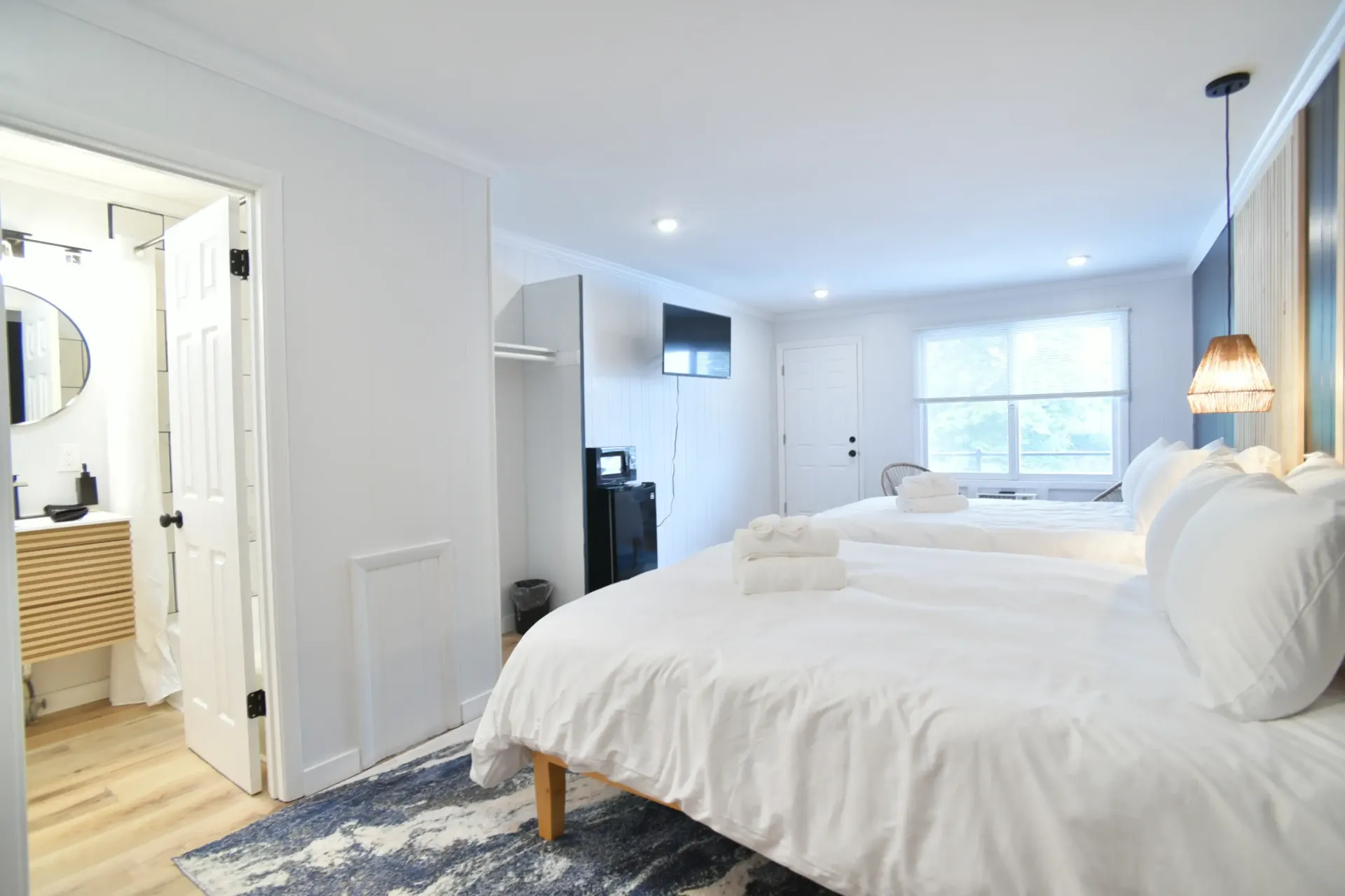 A clean, white hotel room with two beds, a bedside pendant light, a small kitchenette area, and an adjacent bathroom.