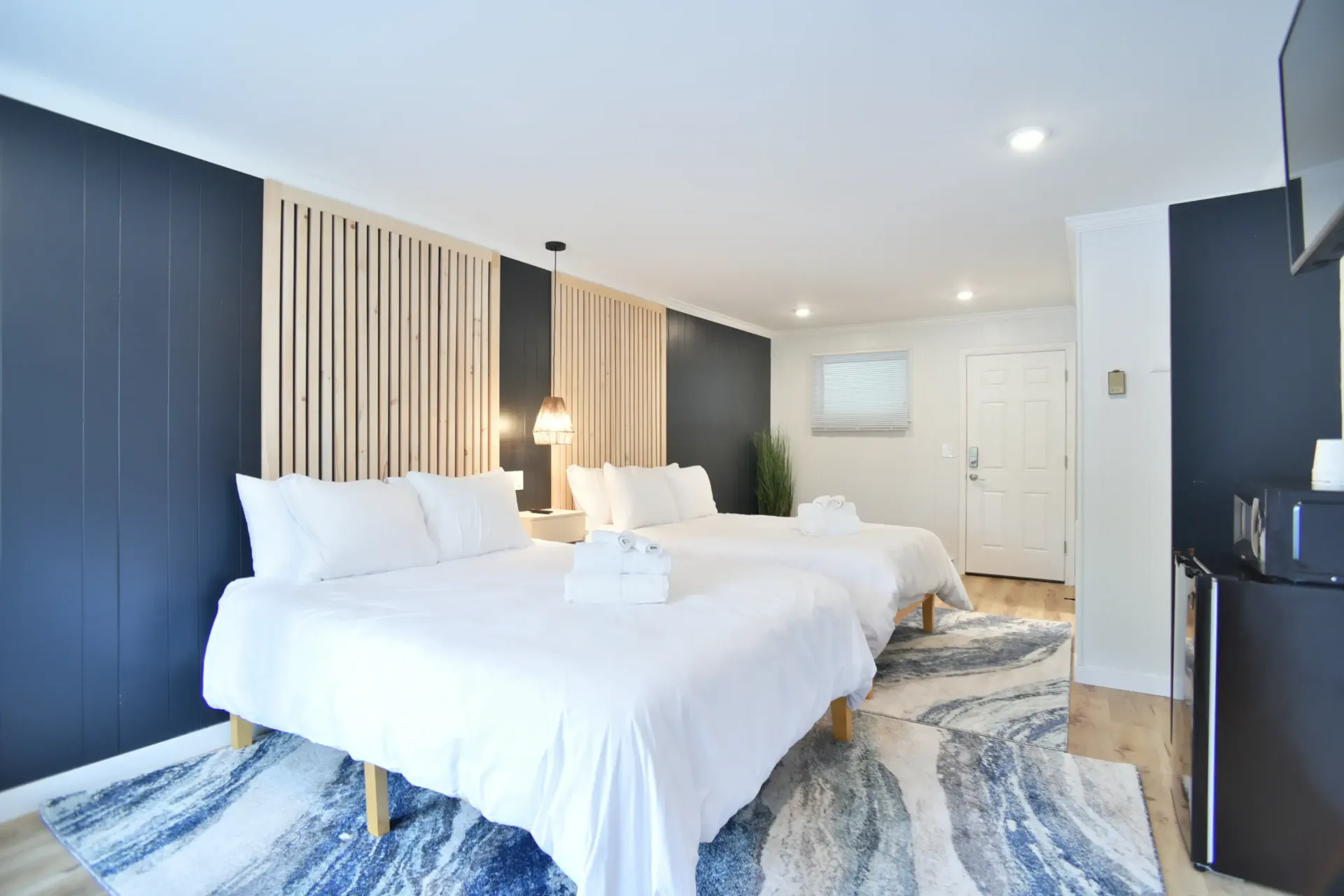 A modern hotel room with two beds featuring white bedding, wood slat headboards, dark blue walls, and a blue patterned rug.
