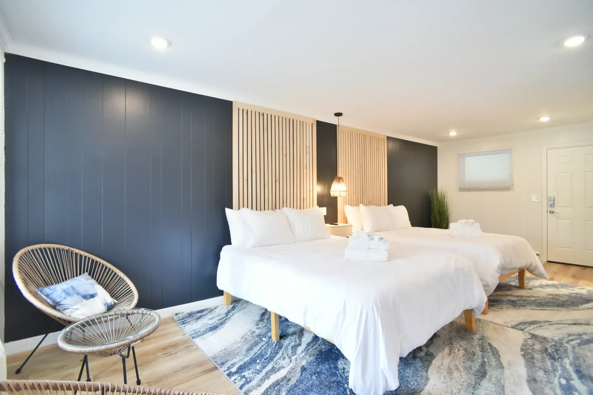 A modern hotel room with two white-made beds, a dark blue accent wall, wooden slats, and a rattan chair on a blue rug.