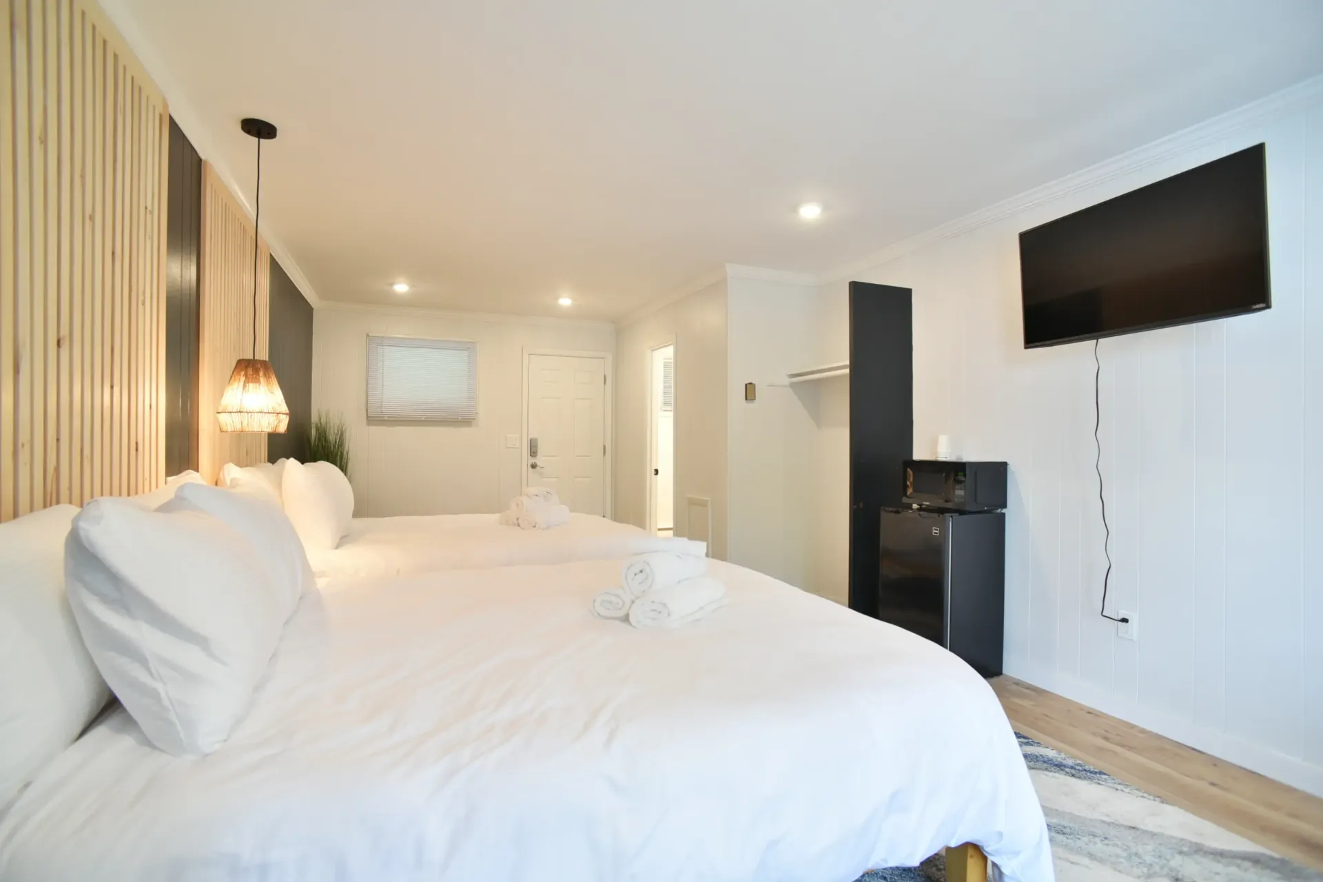 A modern hotel room with two white-made beds, a wall-mounted television, and a small black refrigerator.