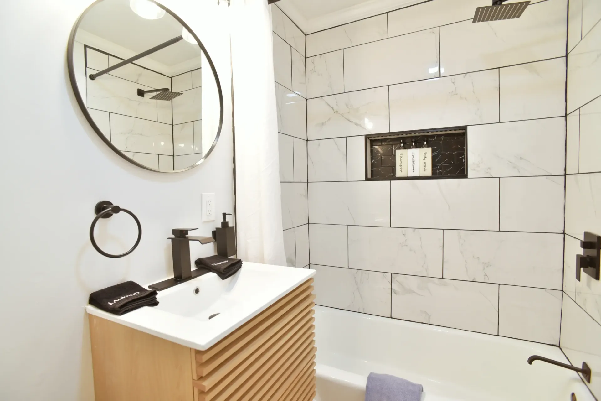 A modern bathroom features a round mirror, light wood vanity, white marble-patterned tiled shower, and dark metal fixtures.