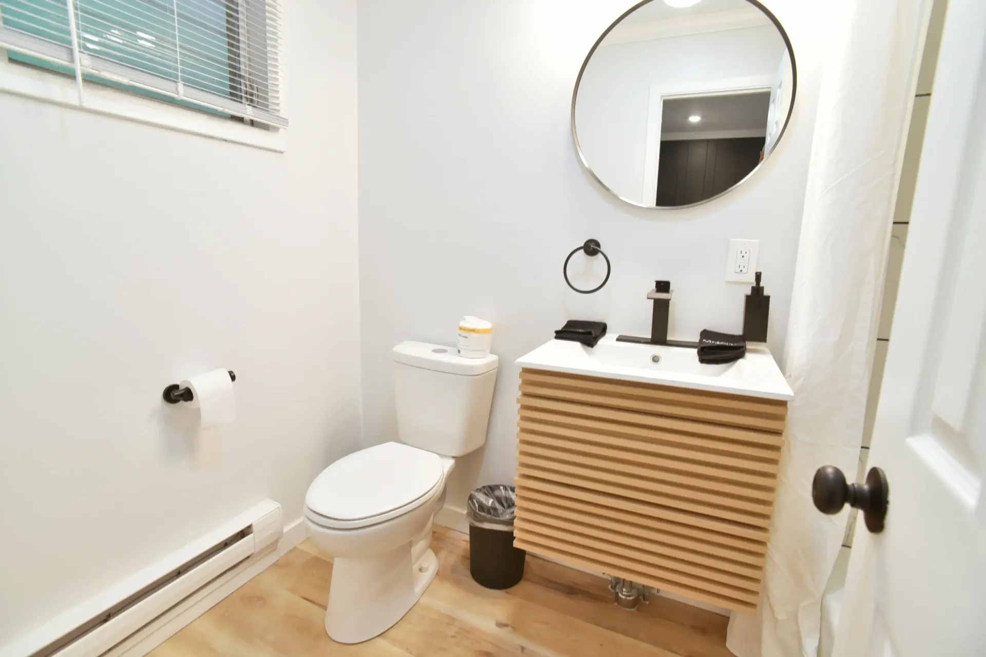 A bright, modern bathroom featuring a white toilet, a slatted wood floating vanity, a round mirror, and a small window.