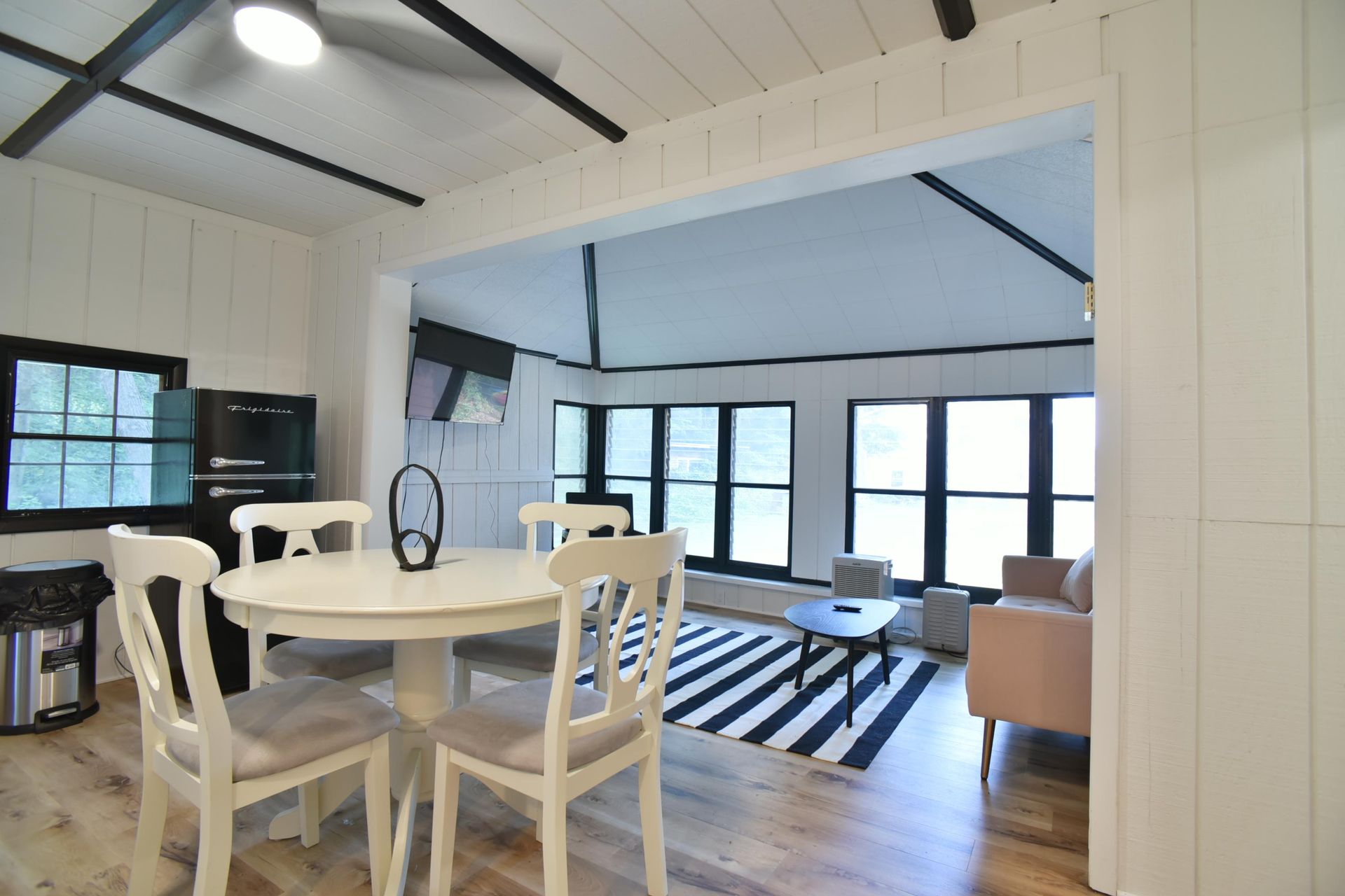 A bright room with a round dining table, four chairs, and a living area featuring large windows and a striped rug.