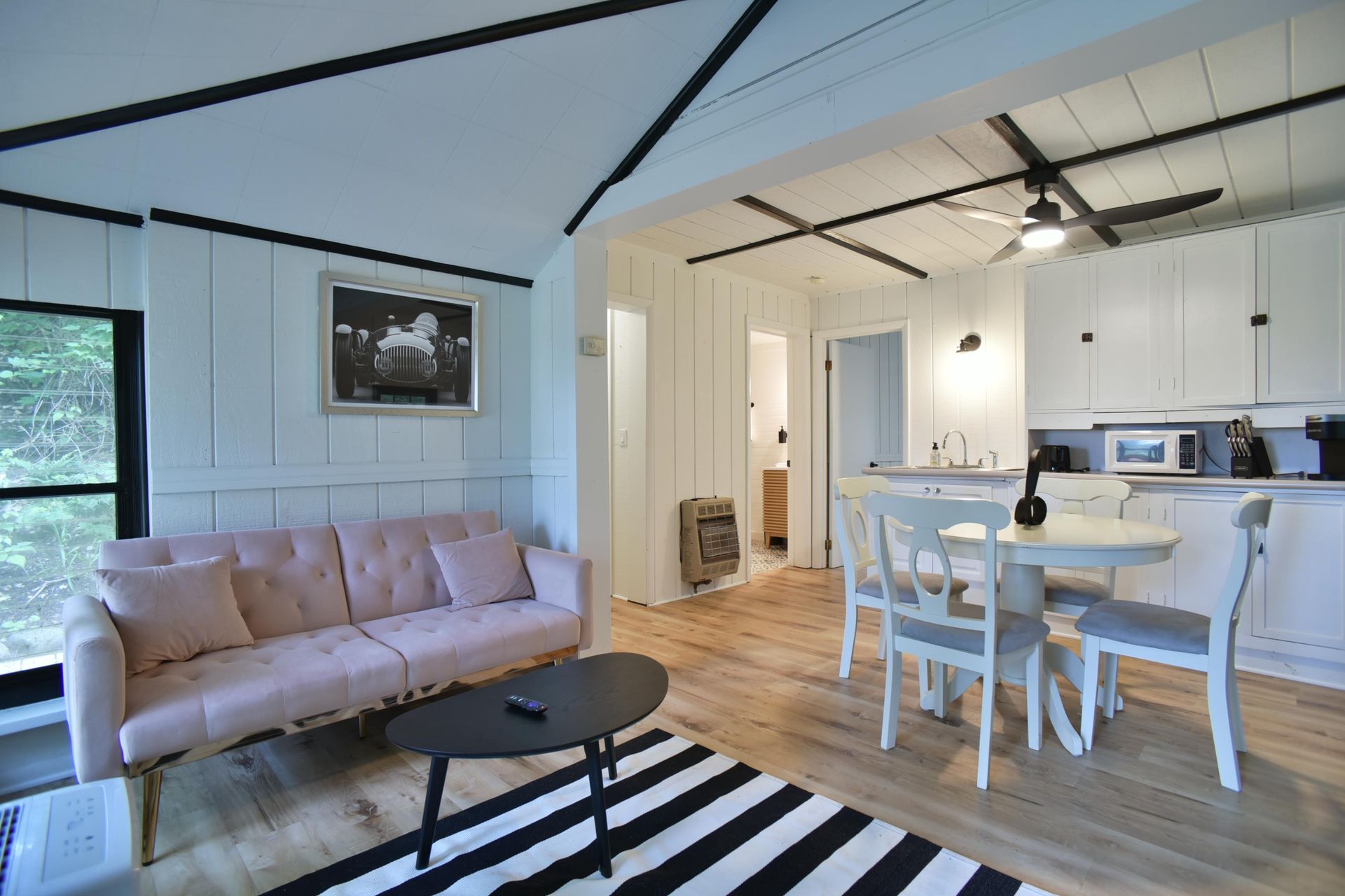 A bright living room and dining area featuring a light pink sofa, a black coffee table, and a small dining set for two.