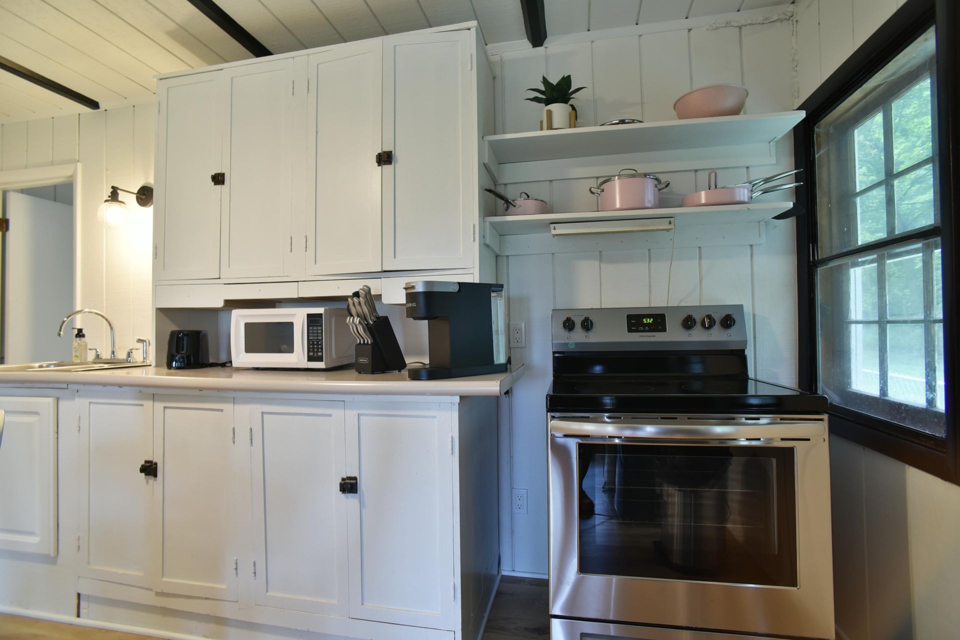 A bright kitchen with white wooden cabinetry, stainless steel appliances, and open shelving holding pink cookware.
