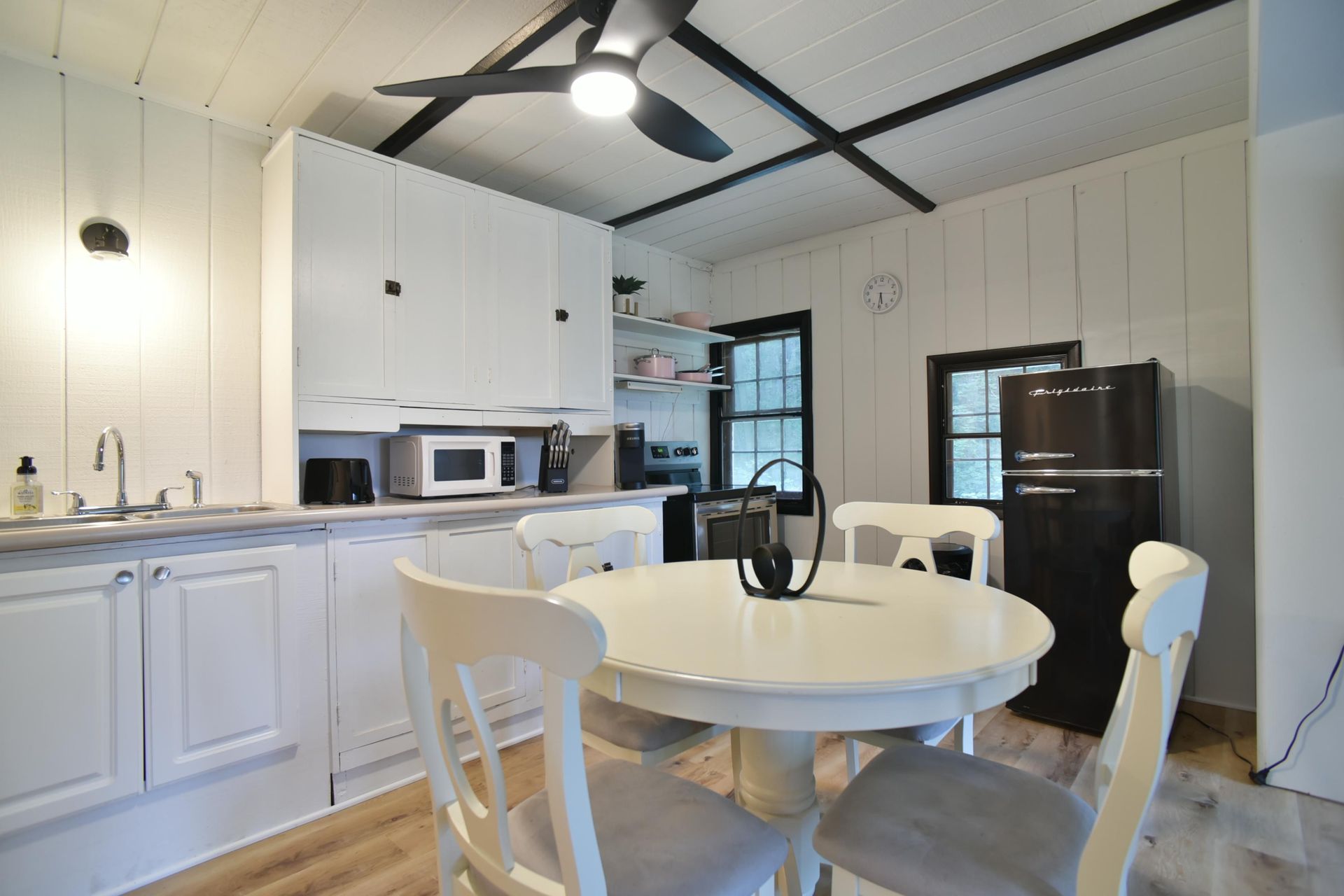 A bright kitchen with white cabinets and wood paneled walls, featuring a round table with chairs and a black refrigerator.