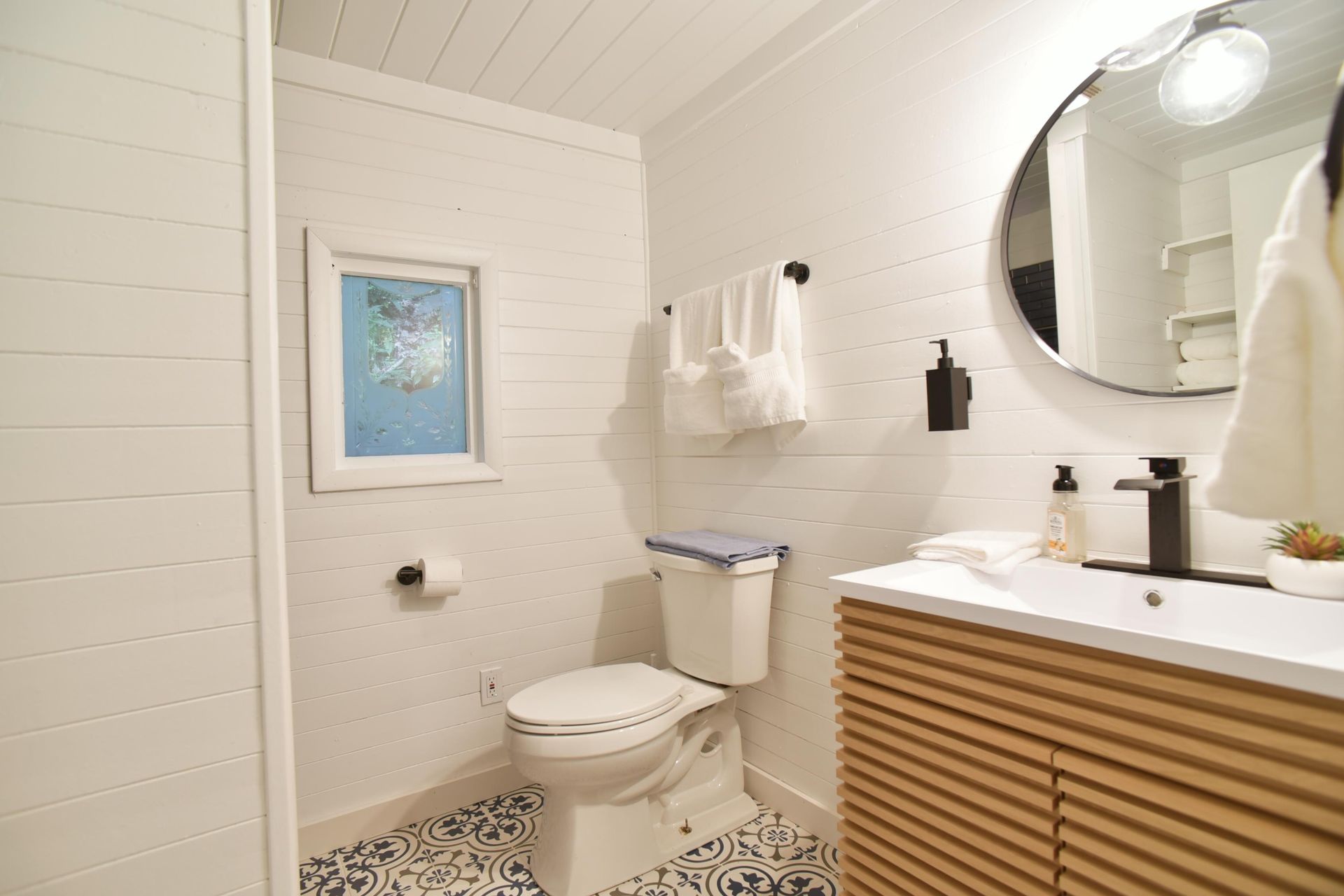 A bathroom with white shiplap walls, a patterned tile floor, a wooden vanity, a round mirror, and a toilet.