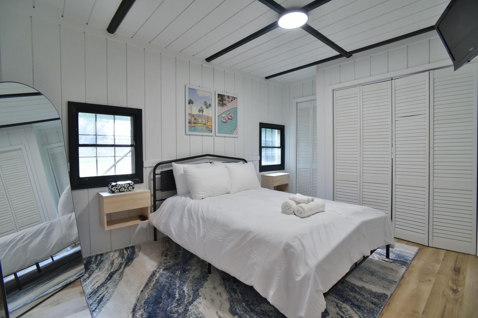 A cozy bedroom with white shiplap walls, a bed, a large arched mirror, and a blue and white abstract area rug.