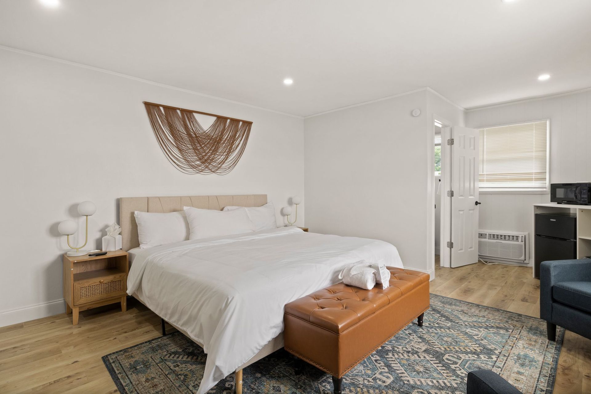 A modern bedroom with a white bed, tan leather bench, patterned rug, macramé wall art, and a side table on wood floors.
