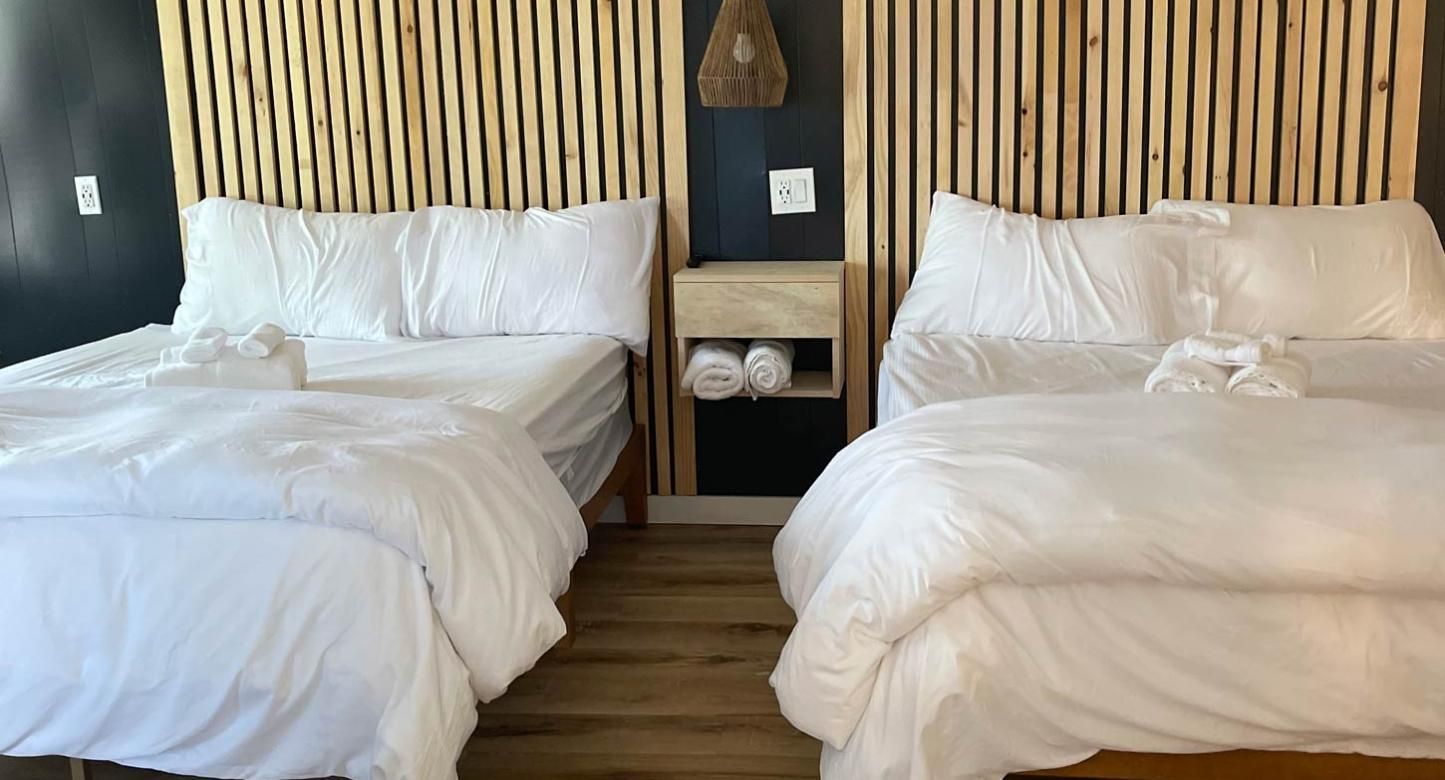 Two white beds with wooden slat headboards and a shared floating nightstand with towels in a room with dark walls.