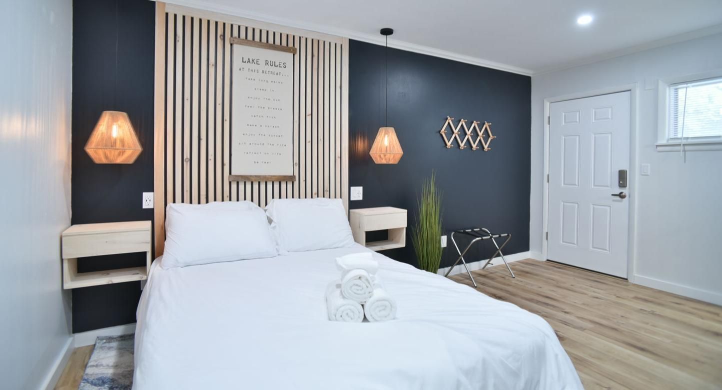 A bedroom with a queen bed, wooden slat headboard, dark accent wall, floating nightstands, and light wood flooring.