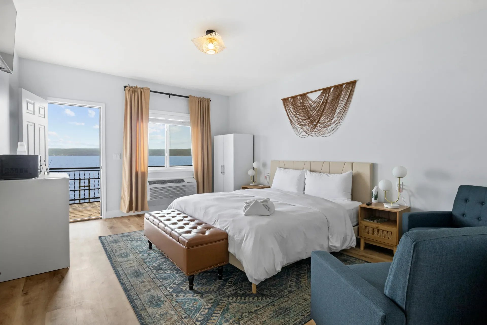 A bedroom with a king bed, patterned rug, brown bench, blue armchair, and a balcony view of the ocean.