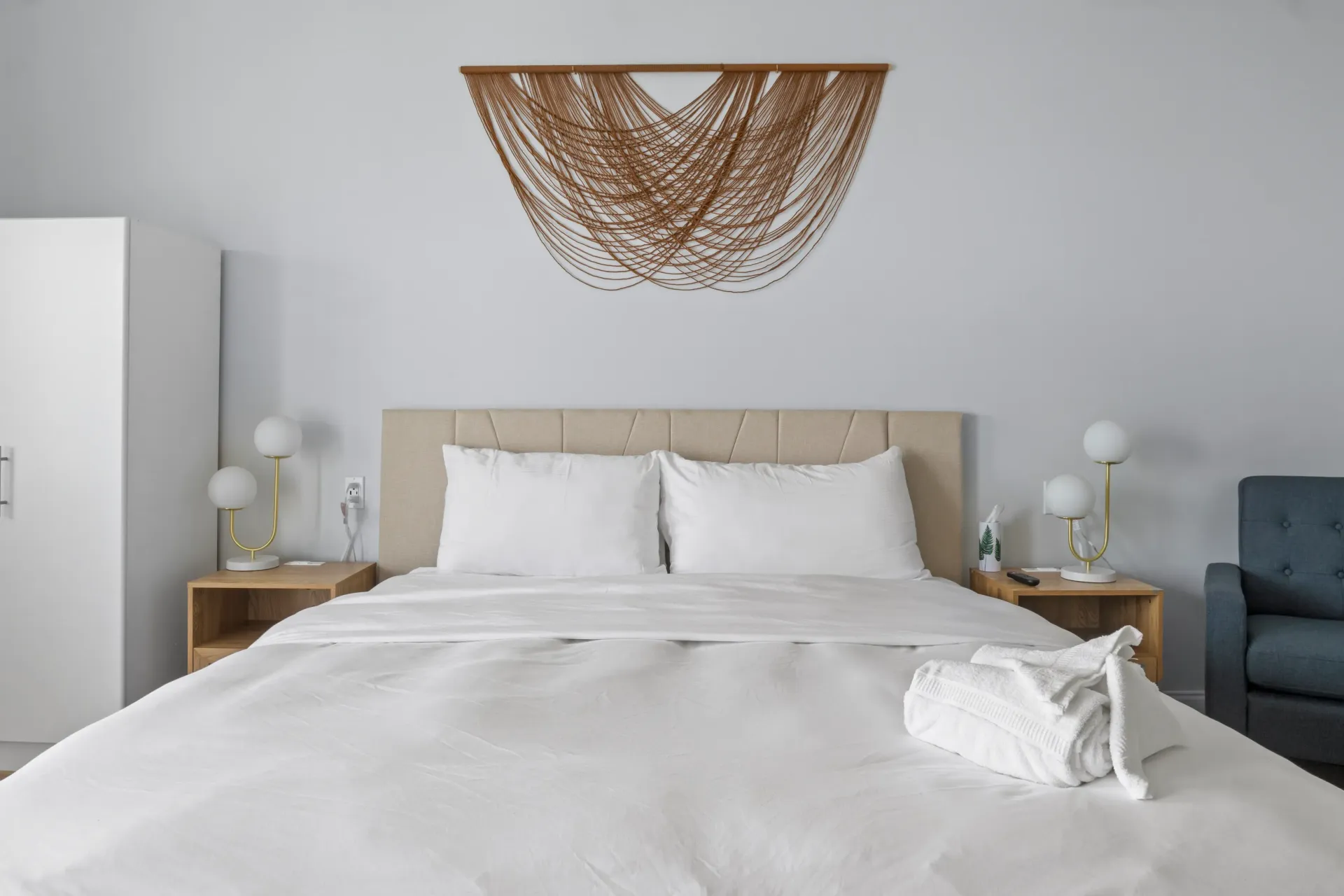 A modern bedroom featuring a bed with white bedding, wooden nightstands, bedside lamps, and a macramé wall hanging.