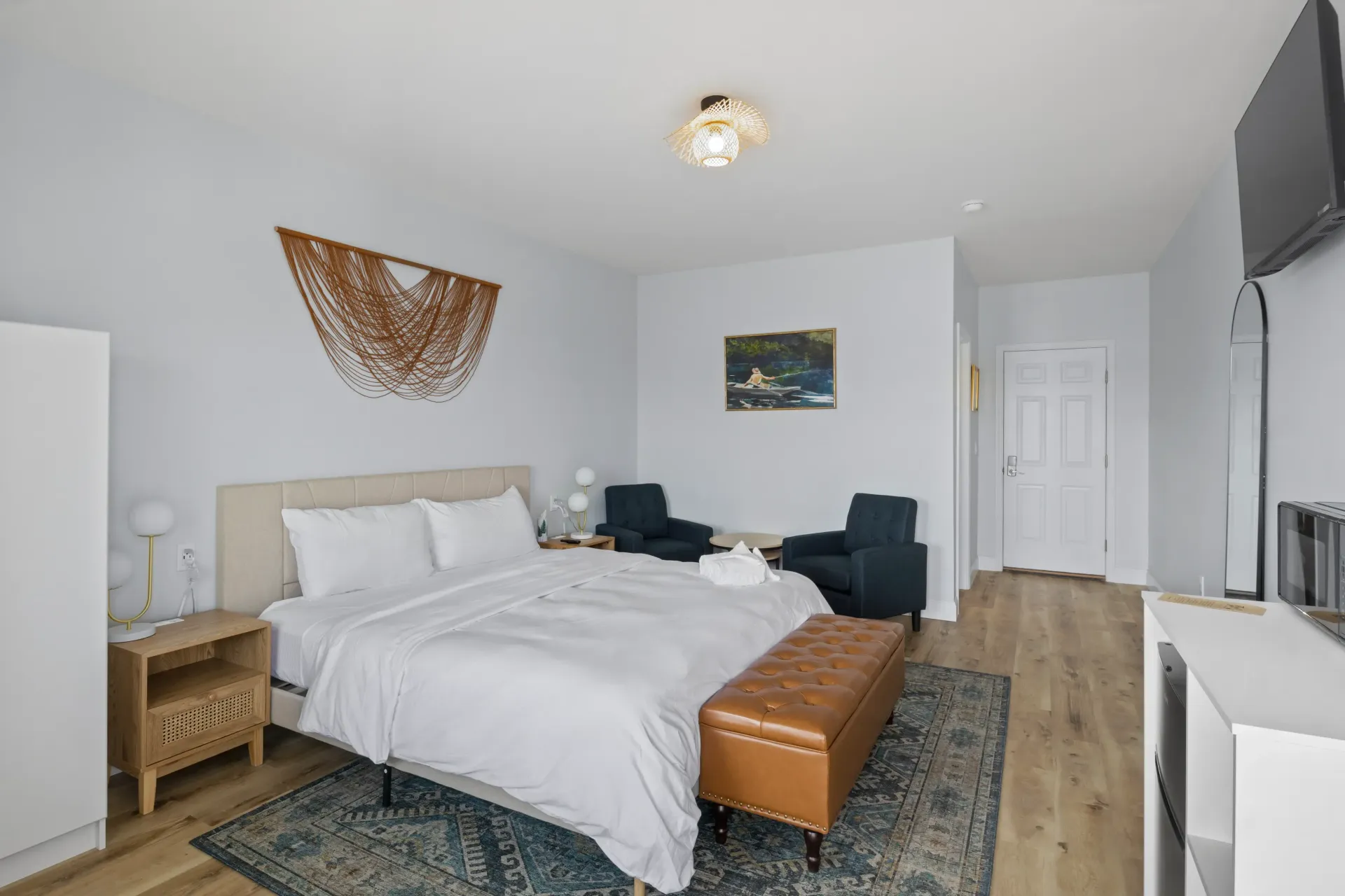 A clean, modern bedroom featuring a bed with white bedding, a brown leather bench, two blue chairs, and wood flooring.