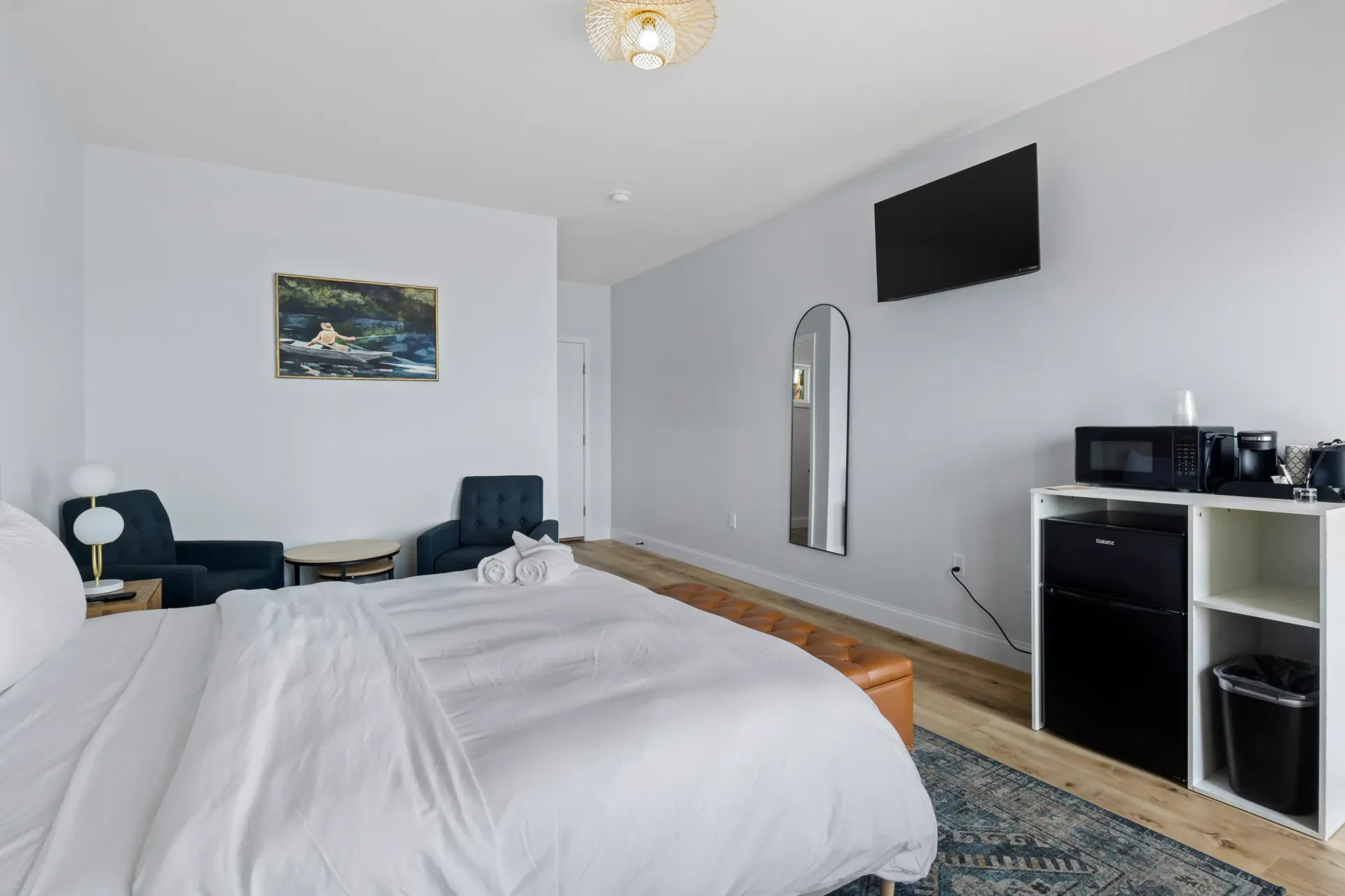 A hotel room featuring a large white bed, two dark armchairs, a television, and a white storage unit with a mini-fridge.