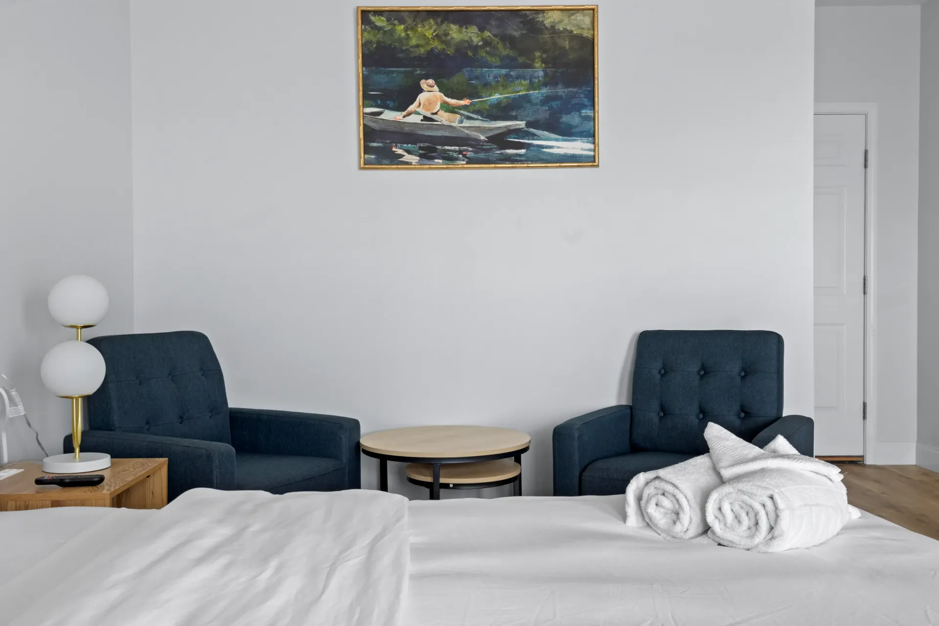 A hotel bedroom featuring a white bed, two blue armchairs, a wooden coffee table, a lamp, and a painting on the wall.