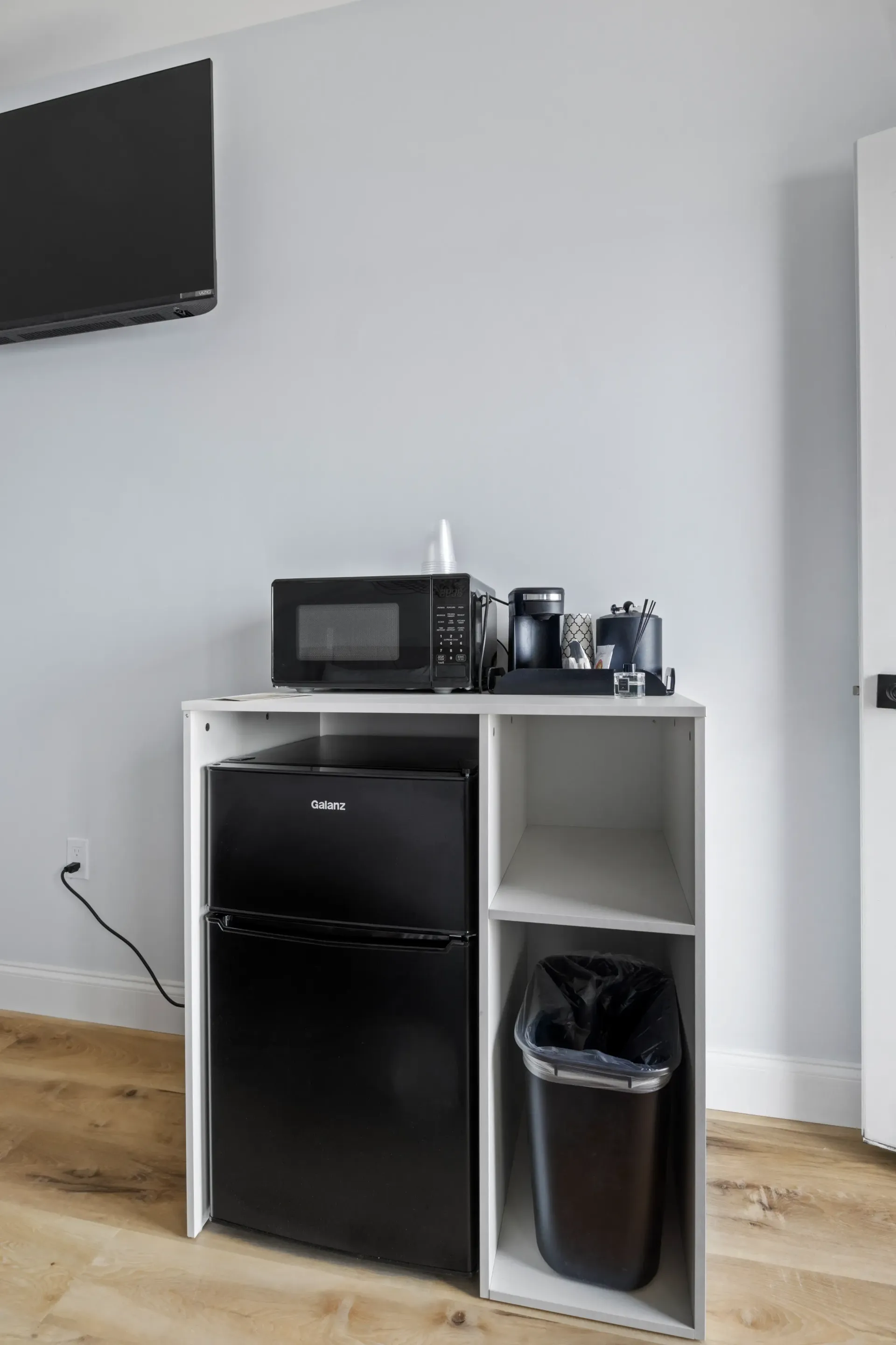 A white cabinet with a mini-fridge, microwave, coffee maker, and trash can against a light grey wall.