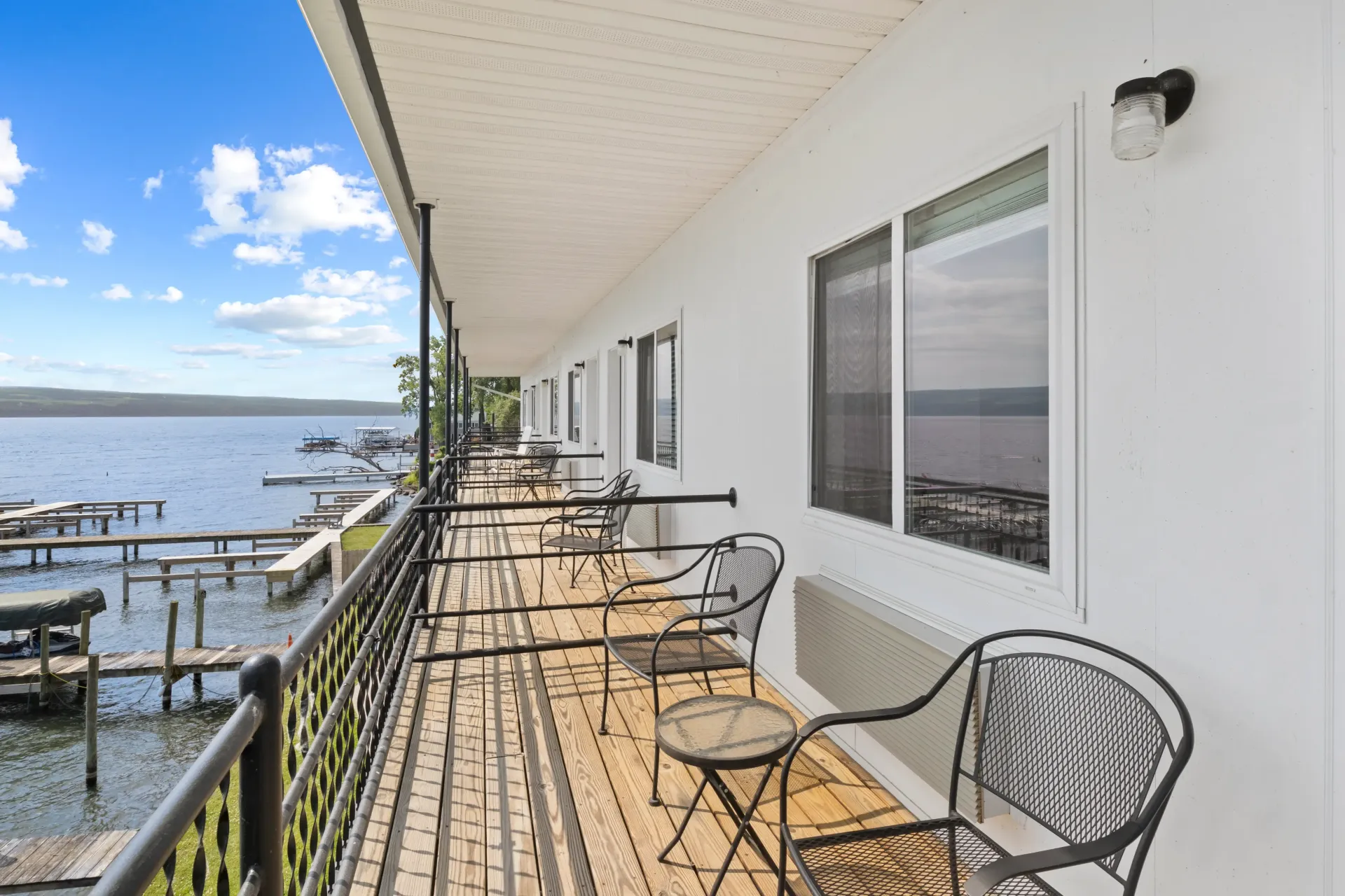 A long, covered outdoor balcony with metal chairs and small tables overlooking a lake with several wooden docks.