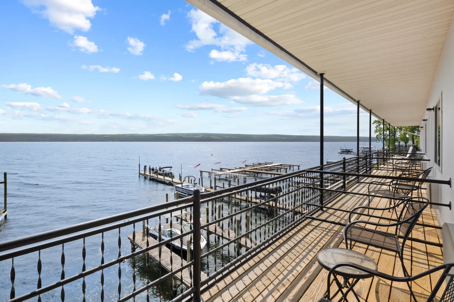 A sunlit balcony with iron railings overlooks a lake with boat docks under a blue sky with scattered clouds.
