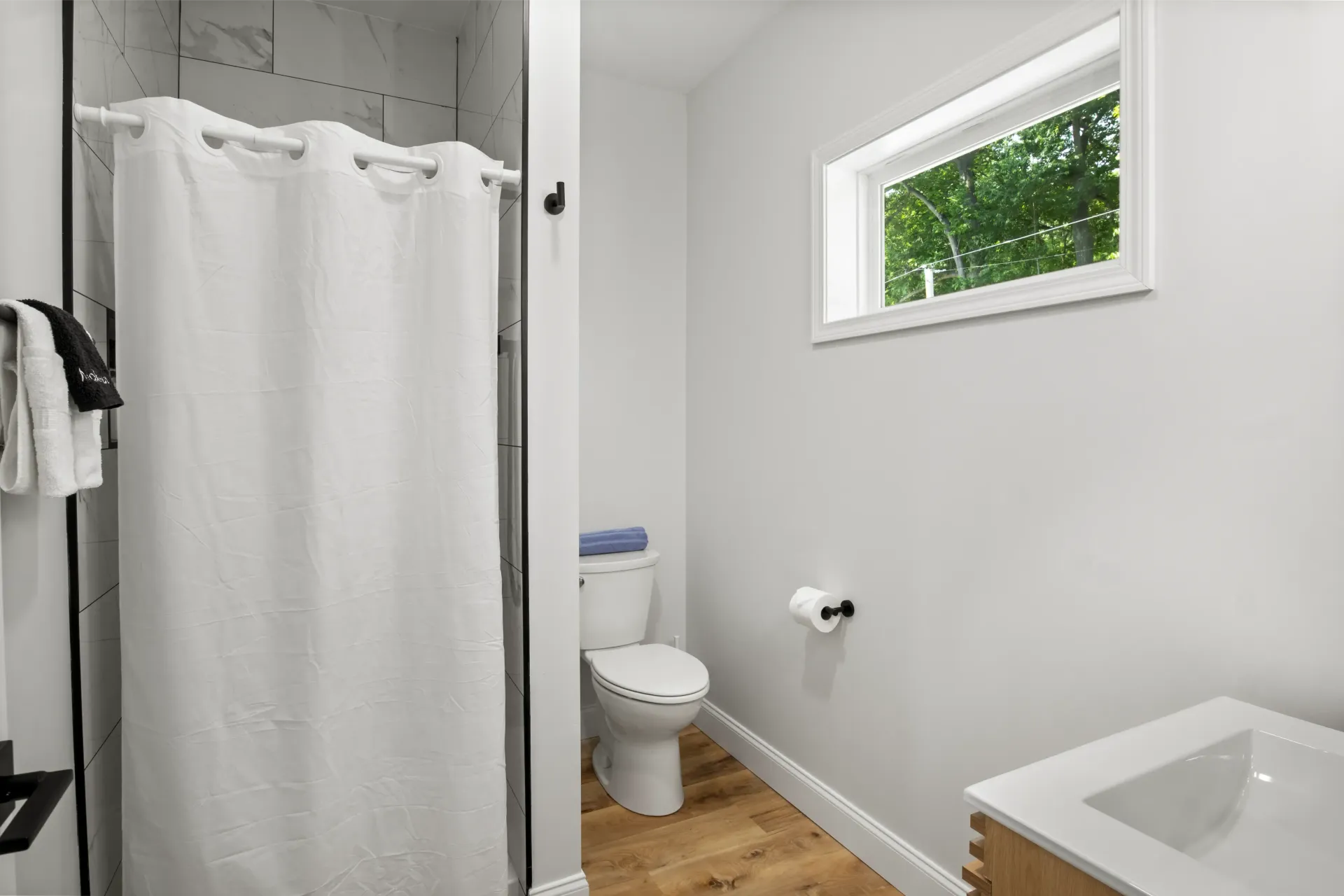 A bright bathroom featuring a white shower curtain, toilet, and a vanity sink under a horizontal window.