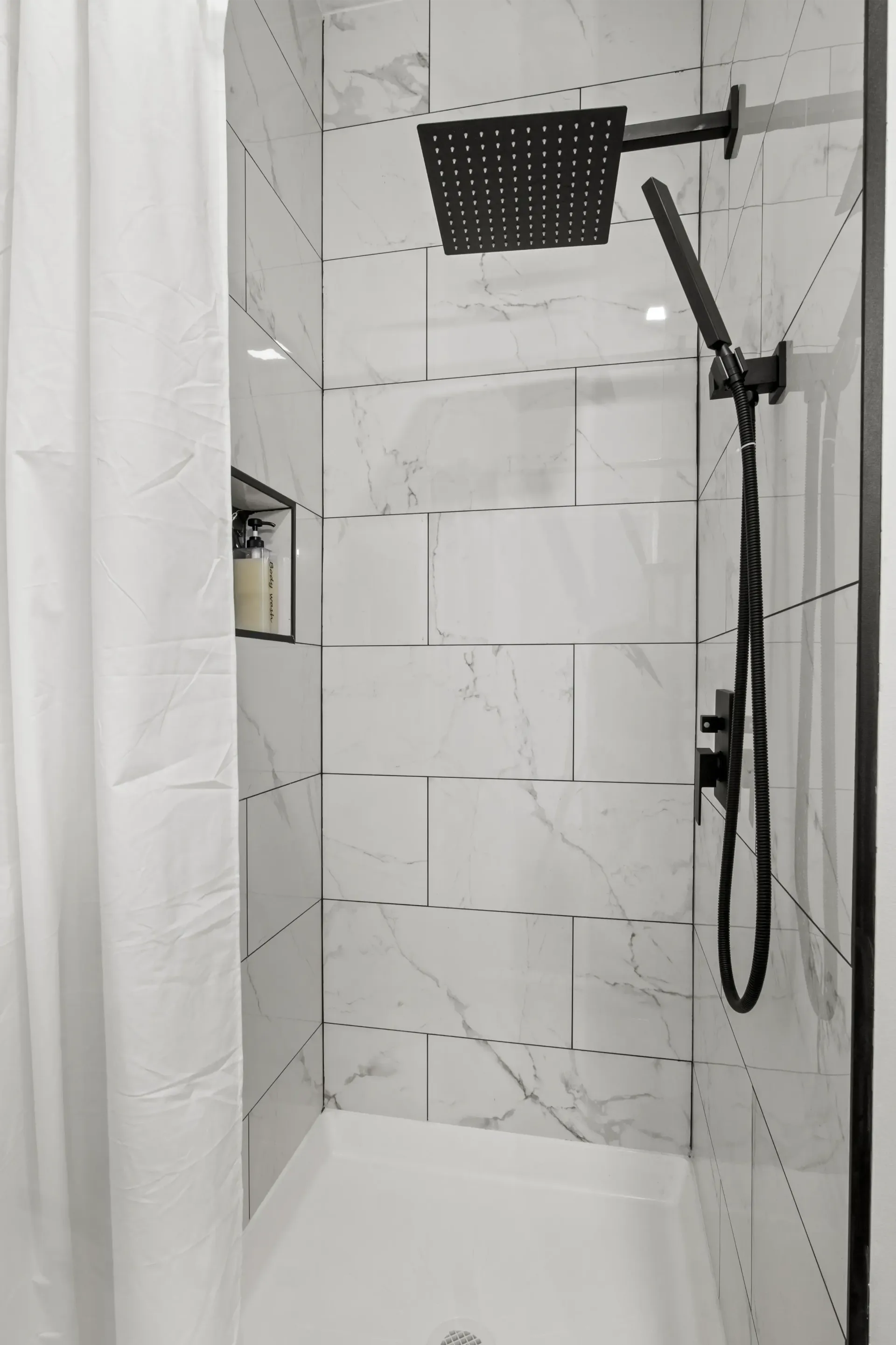 A shower stall with white marble-patterned tiles, a matte black rainfall showerhead, a handheld sprayer, and a niche.