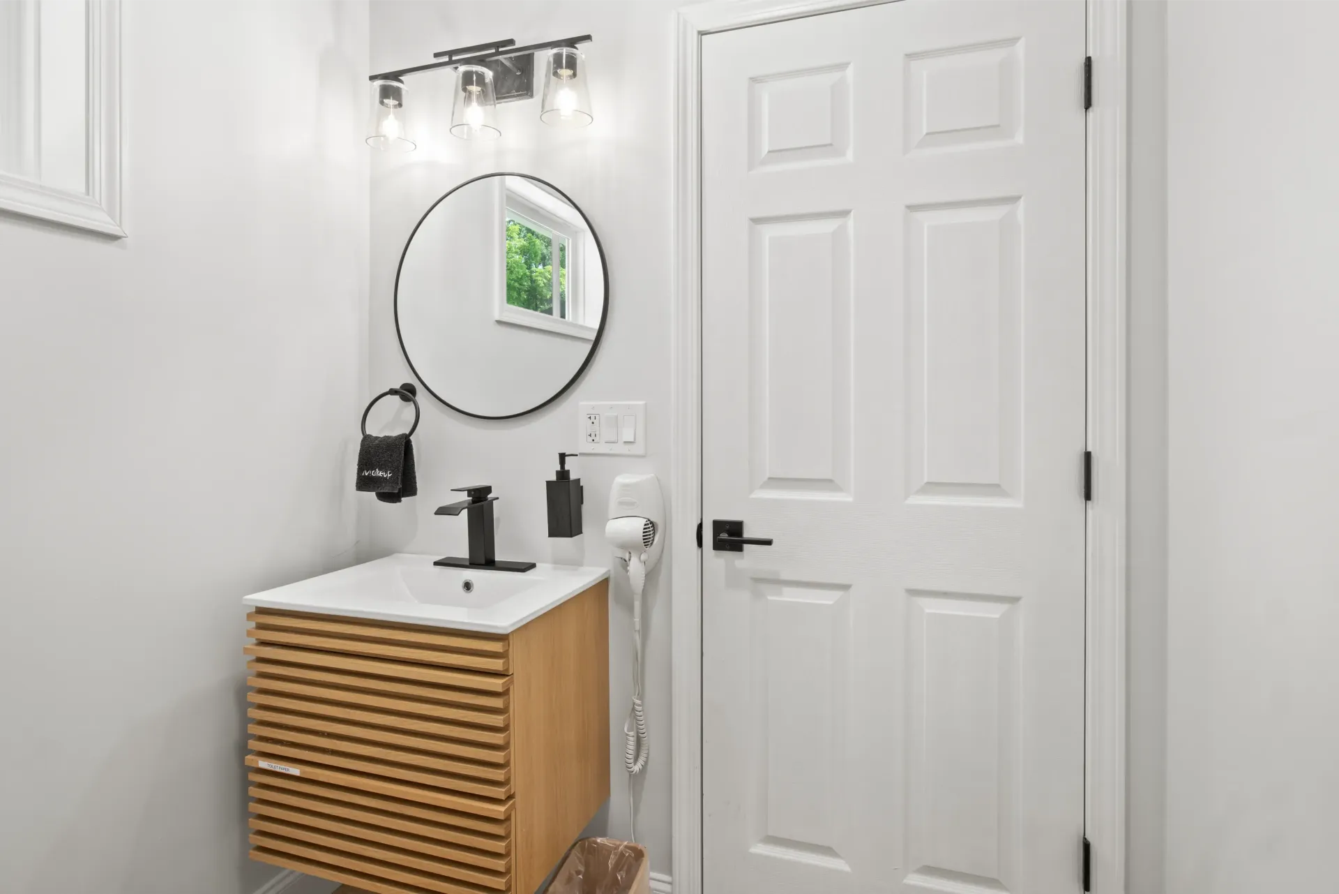 Small bathroom with a wood-slatted vanity, round mirror, black faucet, lighting fixture, and a white six-panel door.