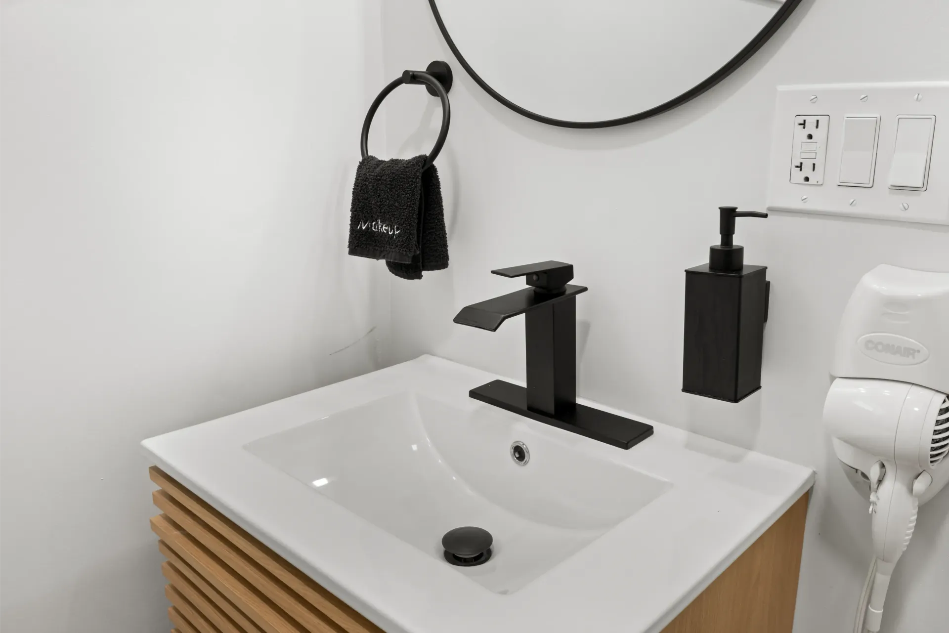 Modern bathroom vanity with a white countertop, black square faucet, round mirror, towel ring, and wall-mounted dispenser.