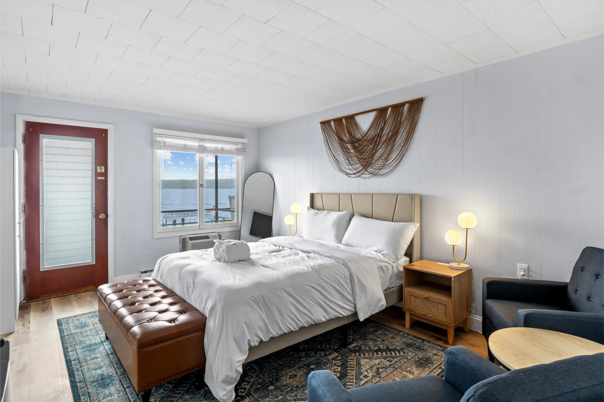 A bedroom with a queen bed, leather bench, and armchairs, featuring large windows overlooking a lake.