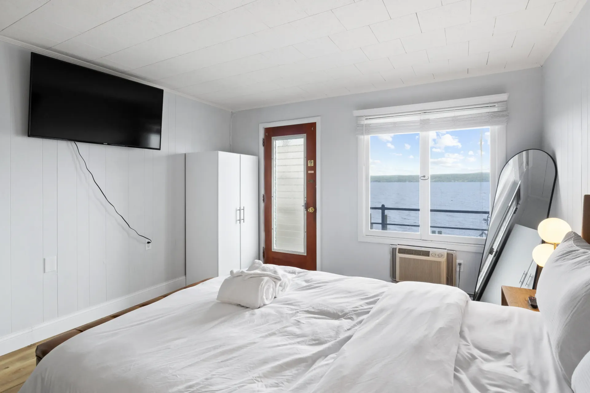 A bright bedroom with a white bed, a wall-mounted TV, a brown door, and a window with a water view.