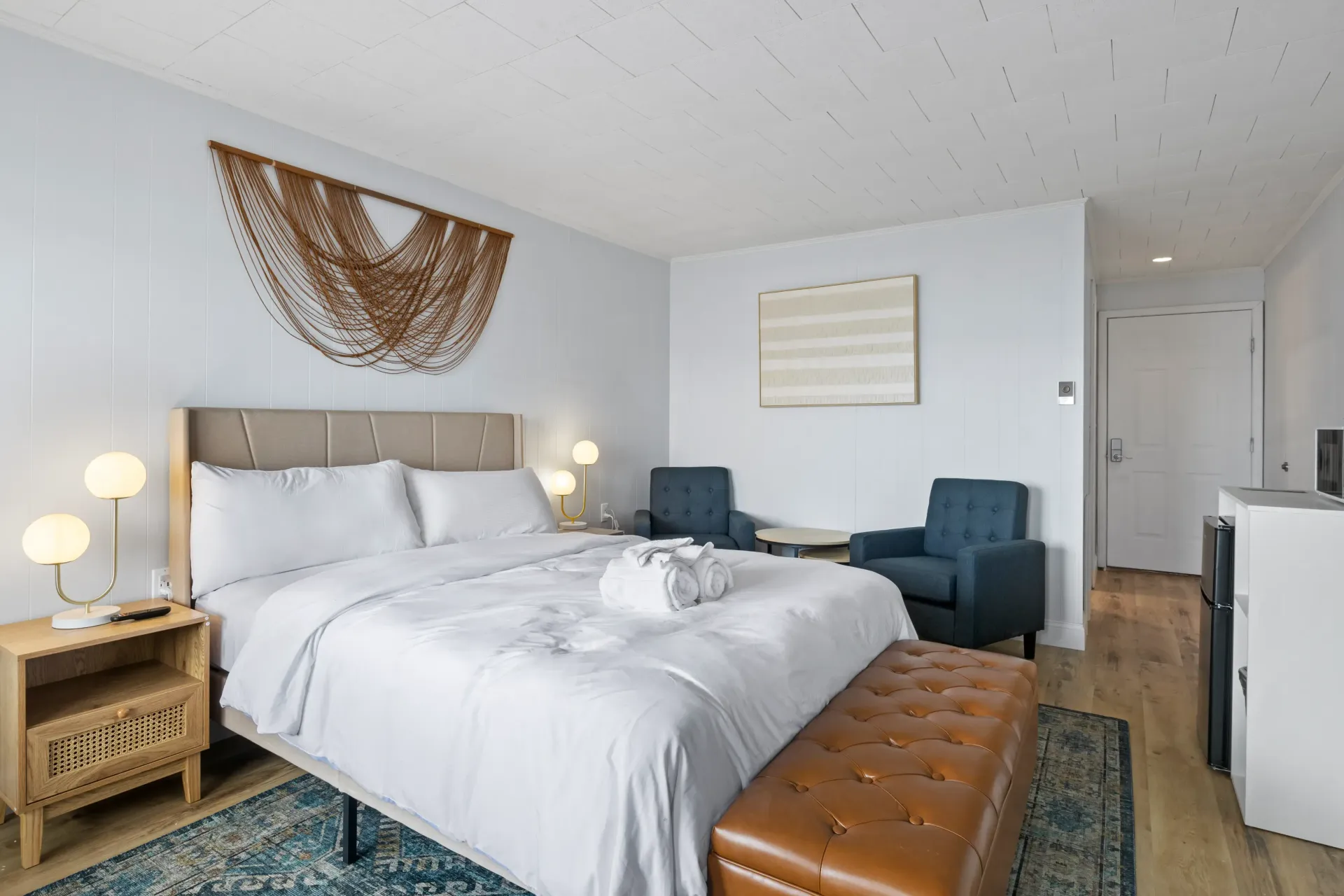 A hotel bedroom with a queen bed, a tan leather bench, two blue chairs, and a textured wall hanging.