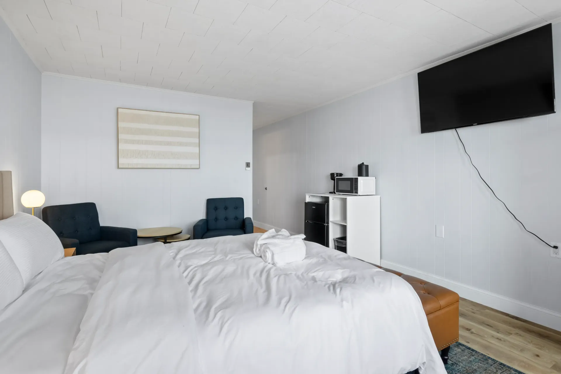 A bright hotel room featuring a bed with white linens, two blue armchairs, a coffee station, and a wall-mounted television.