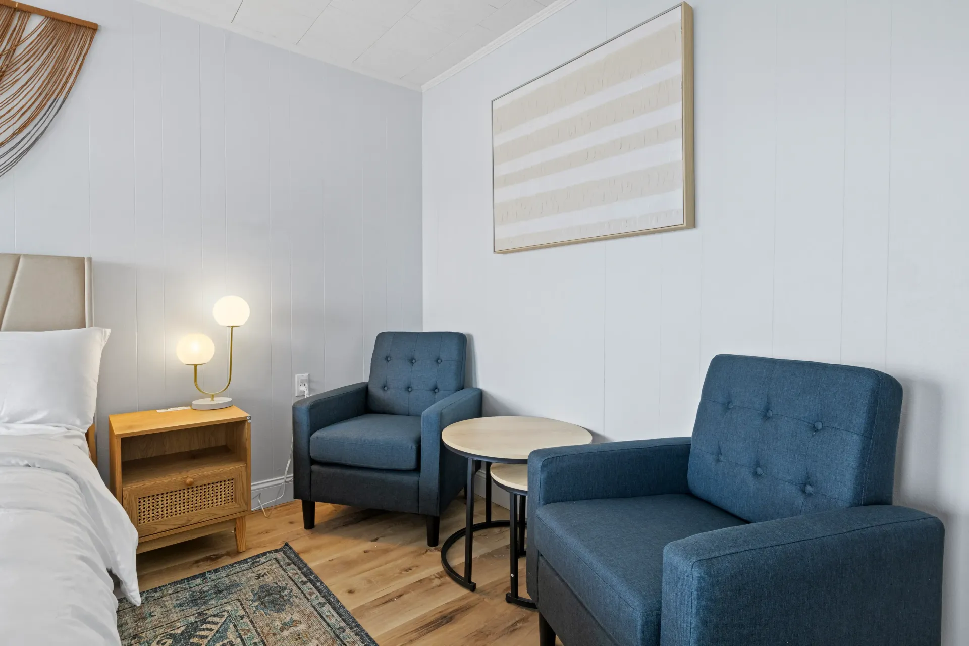 Two blue armchairs in a bedroom, sitting beside a small round table, a wooden nightstand, and a bed with white bedding.