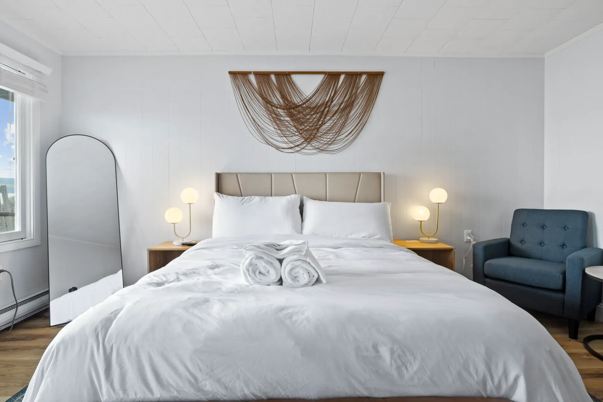 A modern bedroom featuring a bed with white linens, two nightstands with lamps, a blue armchair, and a tall arched mirror.
