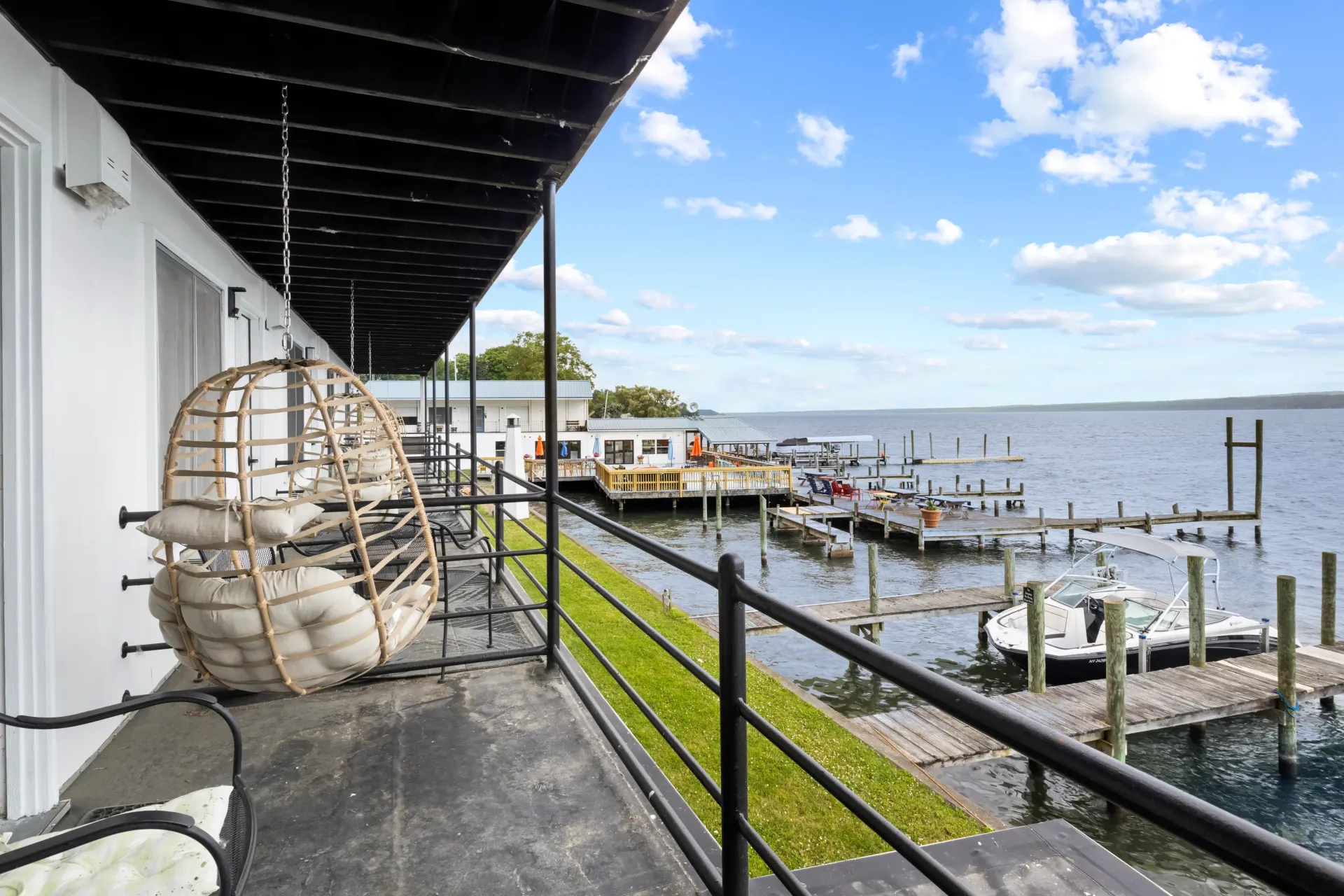 A white balcony with a hanging rattan chair overlooks a canal with docks and boats under a bright, partly cloudy sky.