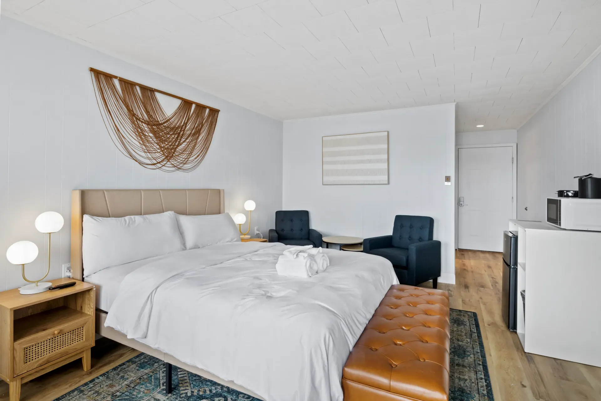 A bright hotel room features a bed with white linens, a tan leather bench, two dark blue chairs, and light wood flooring.